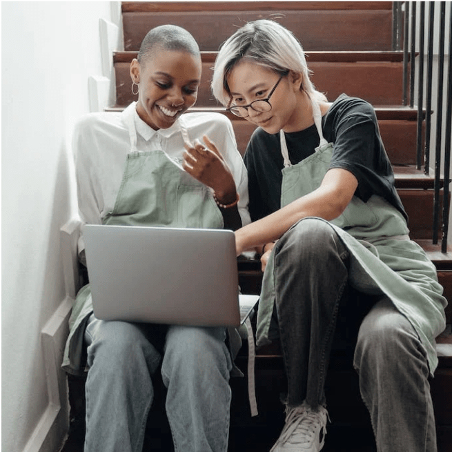 Two women in aprons look at a laptop on stairs.