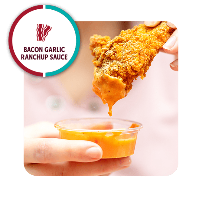A crispy chicken tender dripping with Heinz Remix Bacon Garlic Ranchup Sauce. A hand holds a small cup of the sauce below.