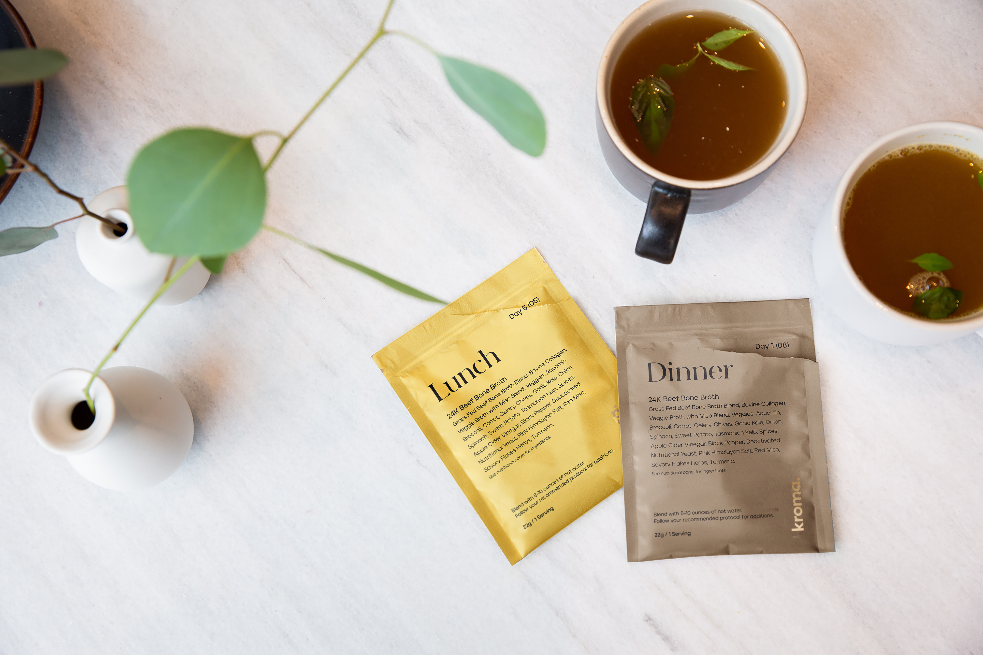 Two 24K Beef Bone Broth sachets next to two mugs of prepared 24K Beef Bone Broth.