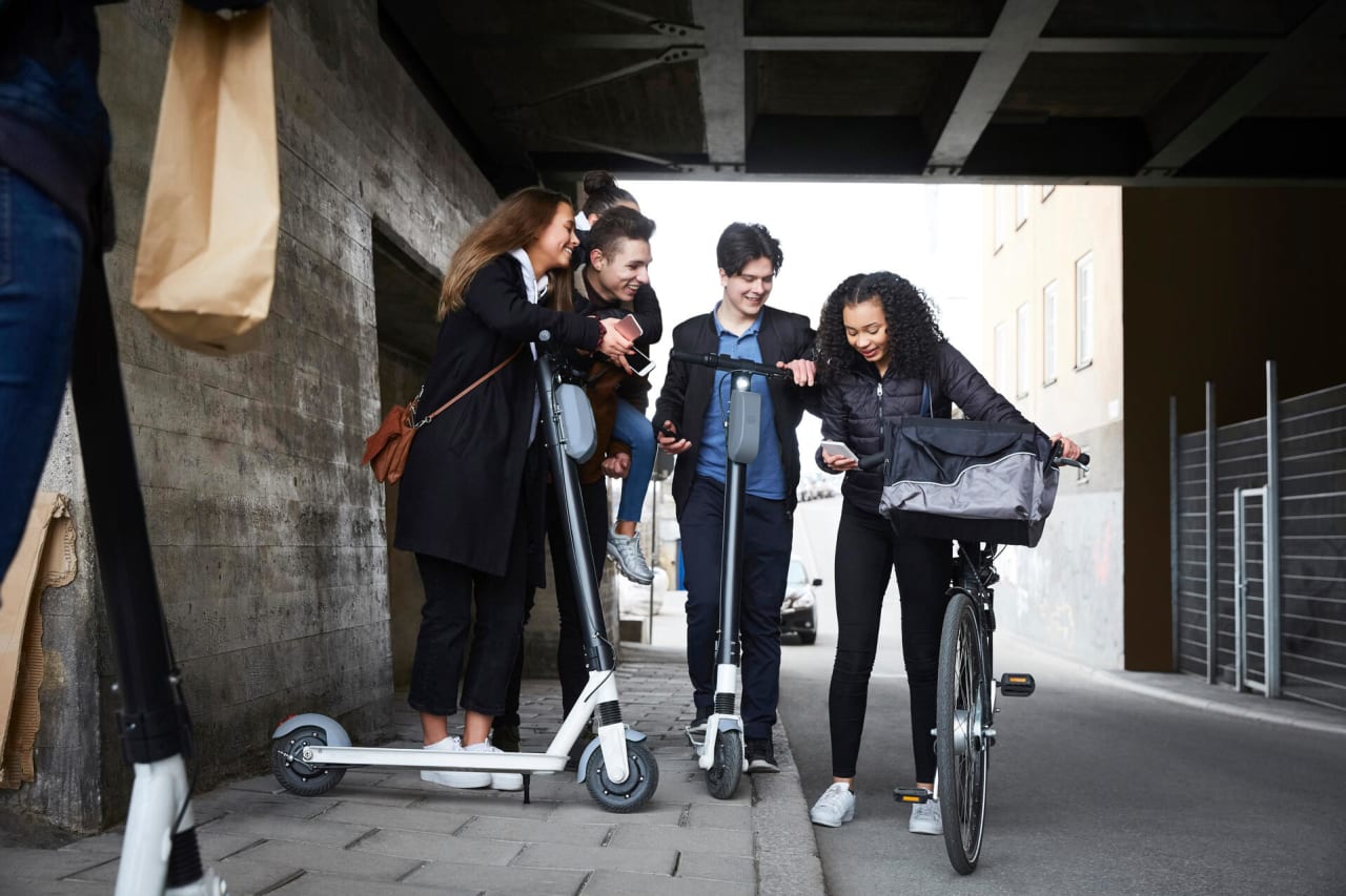 En gruppe leende og glade ungdom i konfirmasjonsalder står i en undergang med sparkesykler og sykler. En av dem holder en mobiltelefon som har alles oppmerksomhet.