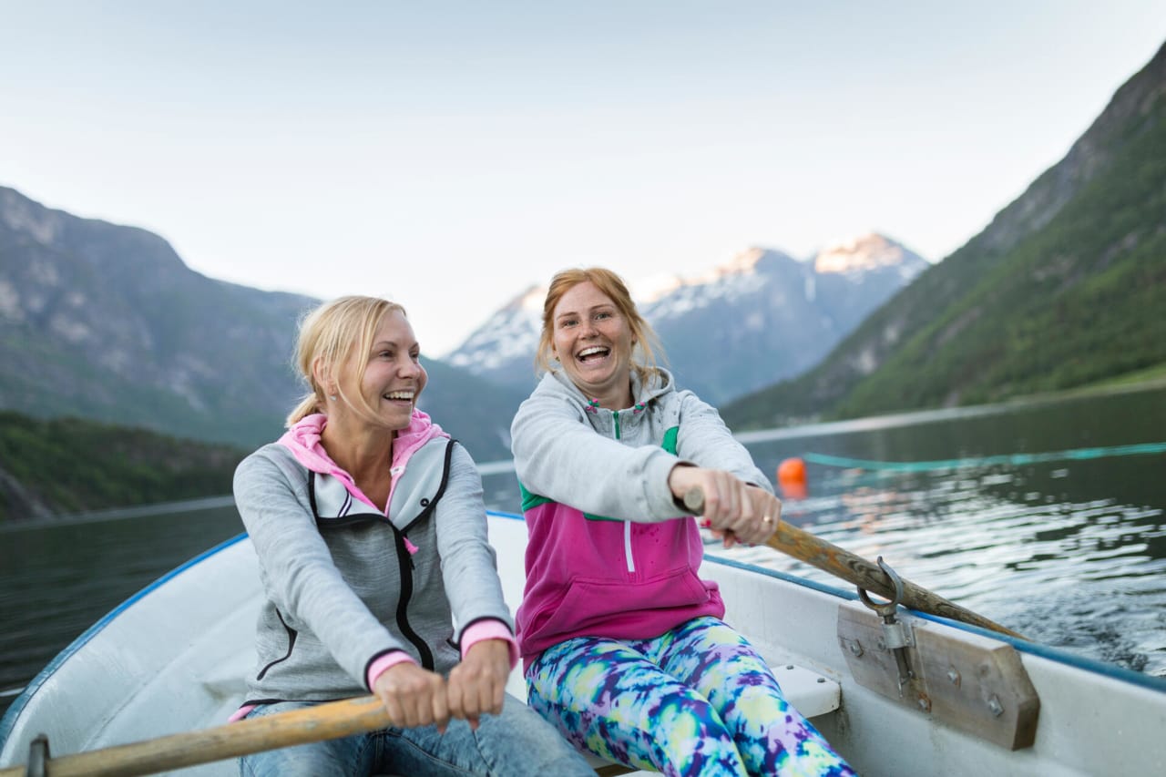 To kvinner holder hver sin åre og ror en robåt, med fjell i bakgrunnen. 