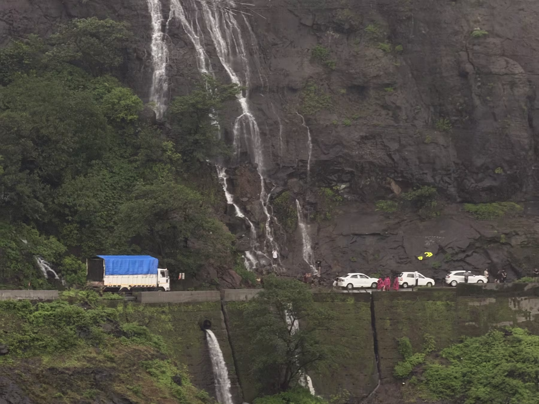 NH61 carved into the raw basalt face: waterfalls crash directly along roadside retaining walls Vehicles passing under roadside rock waterfalls carved into the mountain cliffs of Malshej Ghat