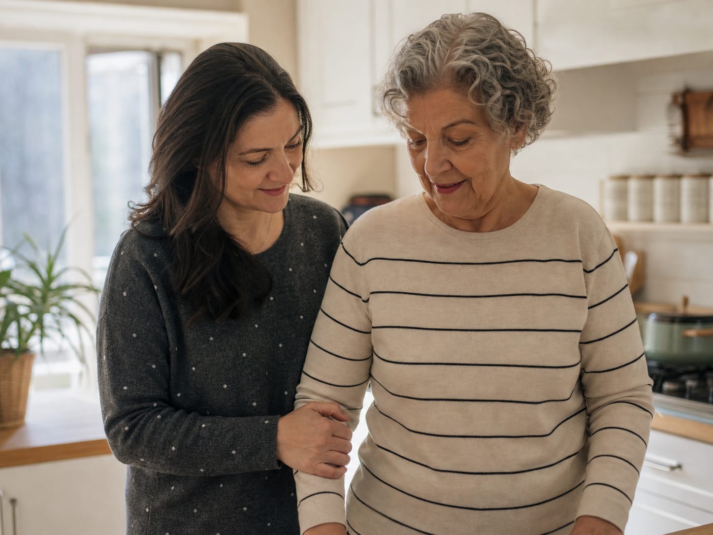 An adult daughter supporting her older mother in a home kitchen.