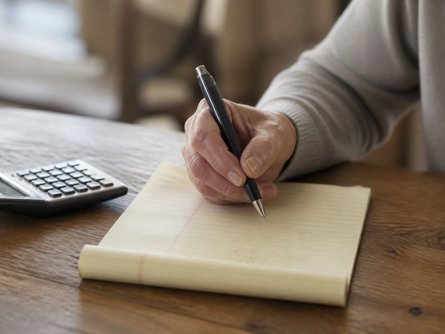A hand making a household-cost list on a legal pad beside a calculator.