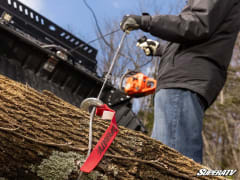 Winch rope and red strap wrapped around a log, with a man holding a chainsaw in the background.