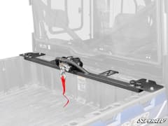 Black winch crossbar mounted across the front of a UTV bed, behind the cab, with a red hook strap hanging down.