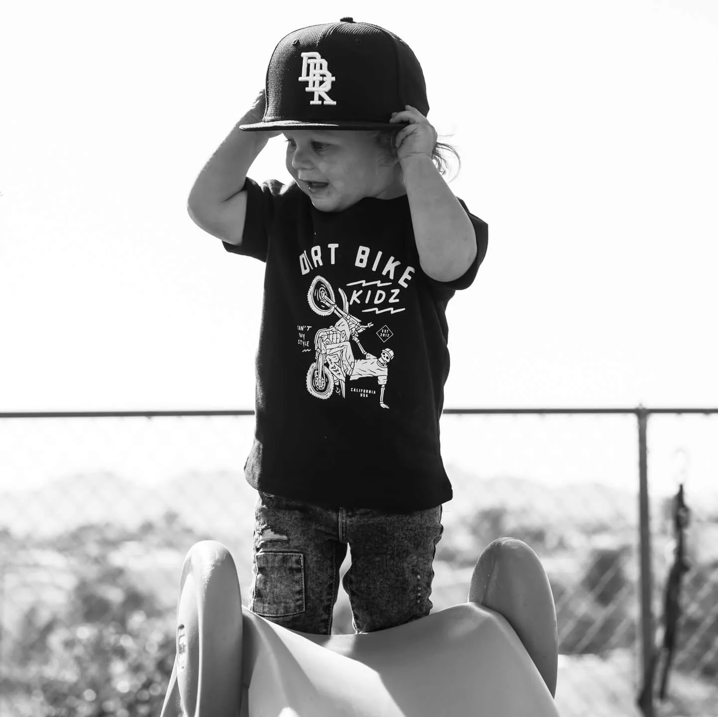 Toddler wearing the DBK 12 O' Clock Toddler Tee and a DBK cap outdoors, black and white photo