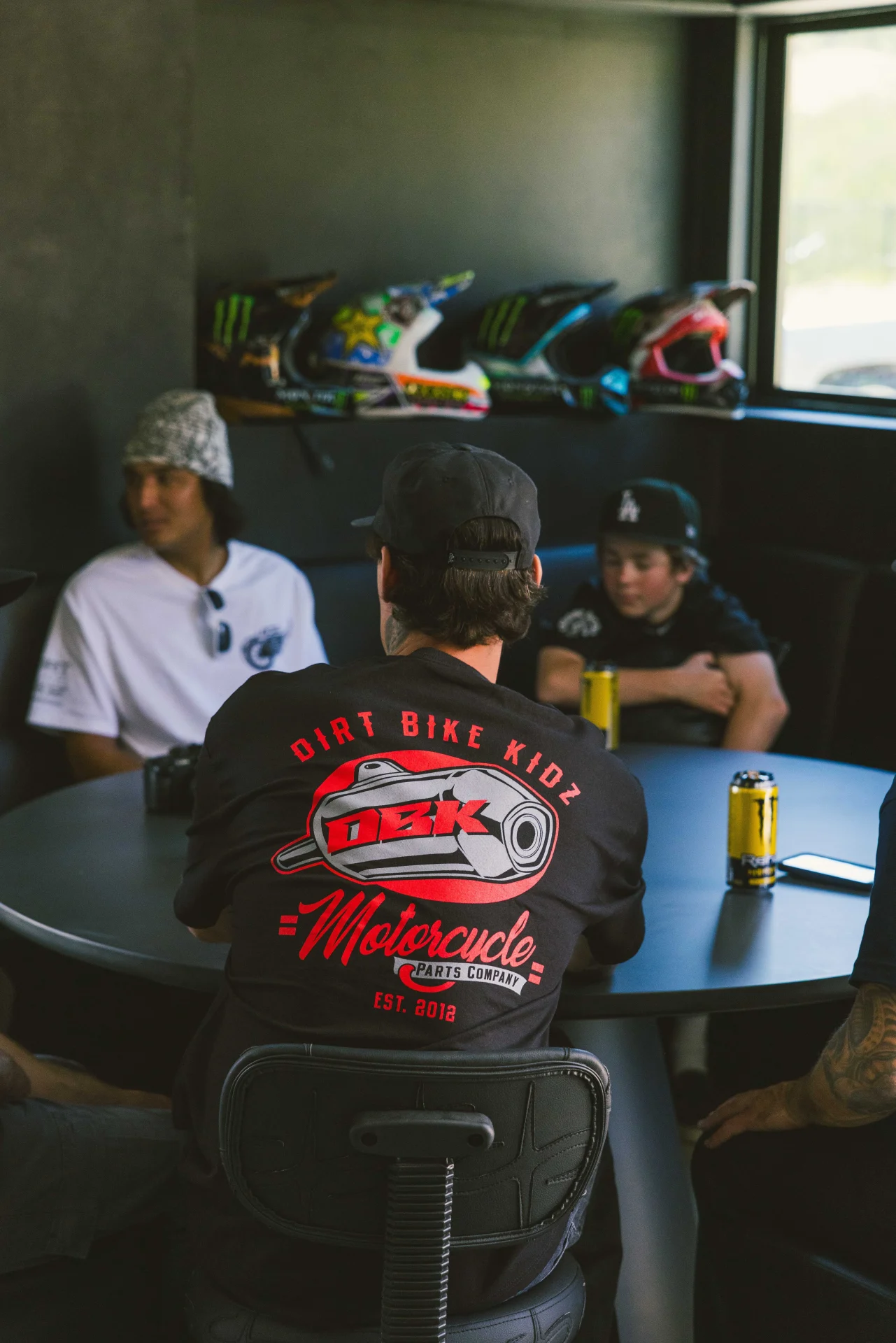 Person wearing the black DBK Factory tee seated at a shop table, back graphic visible