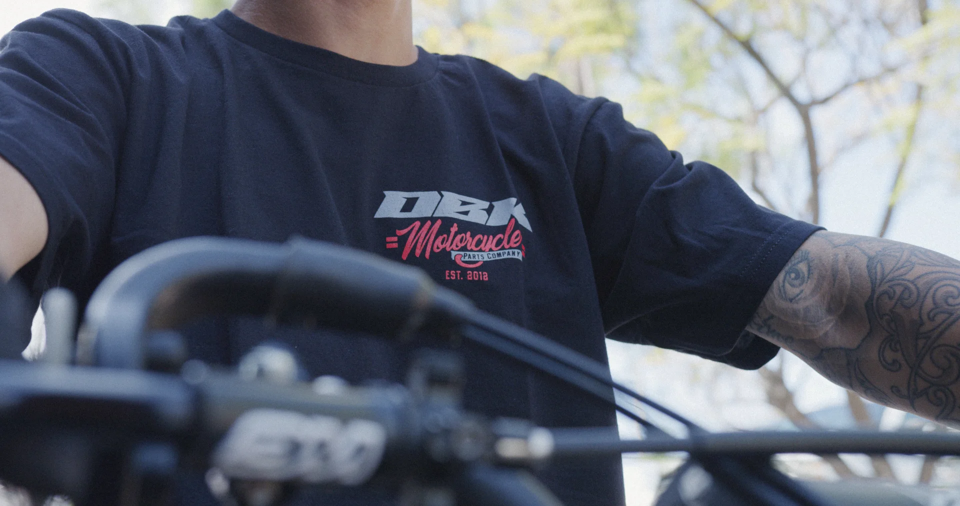 Rider wearing the black DBK Factory tee behind dirt bike handlebars, chest logo visible