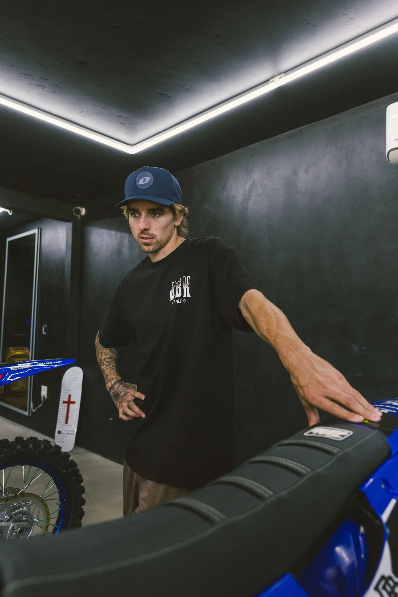 Rider wearing the DBK Ghost Rider Tee in black, leaning on a dirt bike in a garage