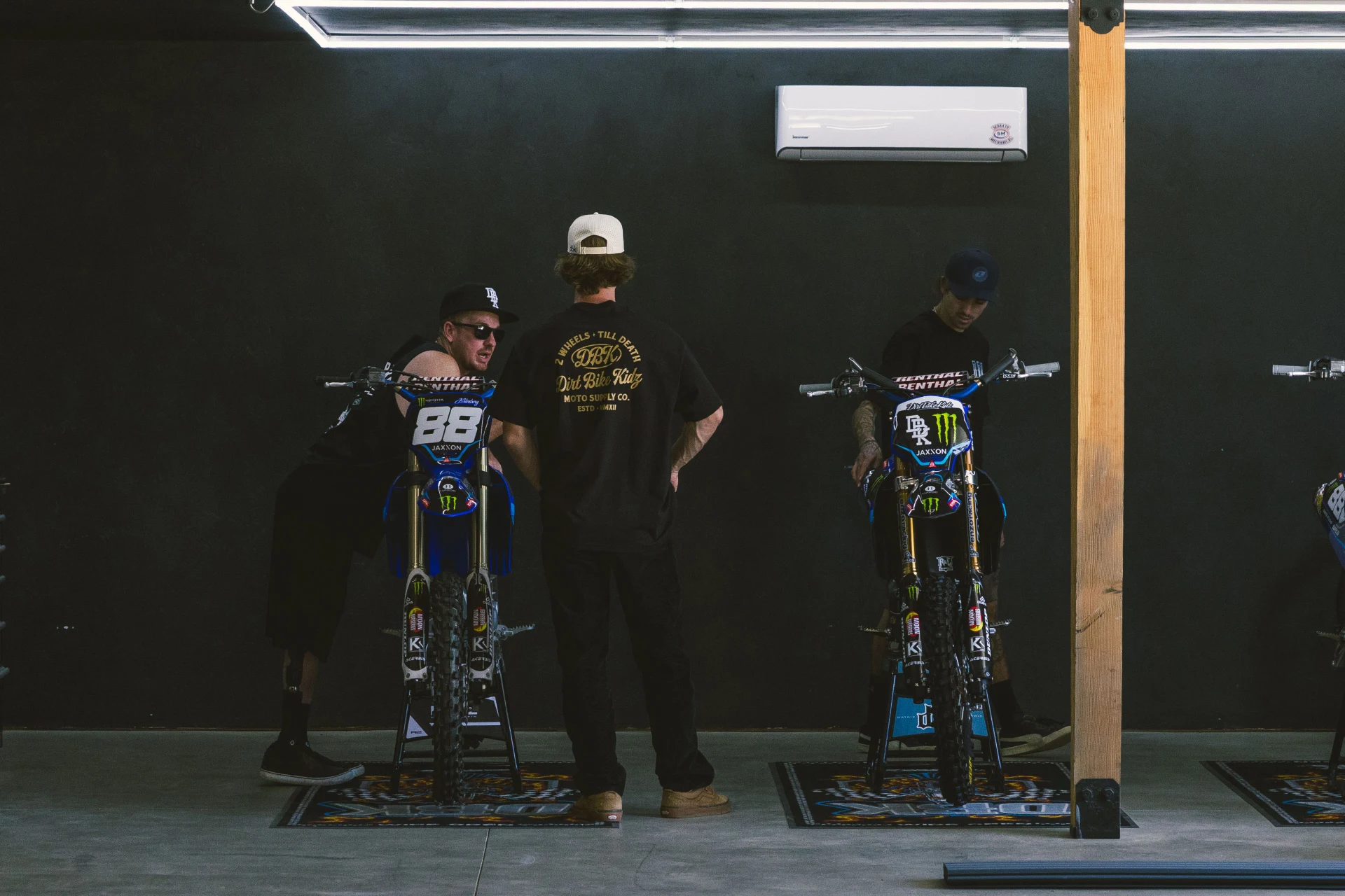 Two people in a garage with dirt bikes, one wearing the DBK Gold Nugget Tee in black