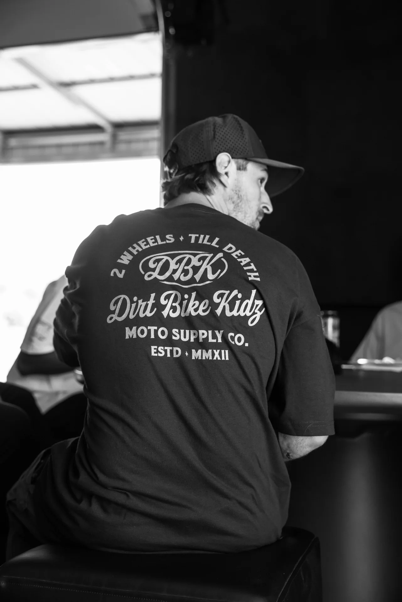 Black and white photo of a man seated in the DBK Gold Nugget Tee, back view