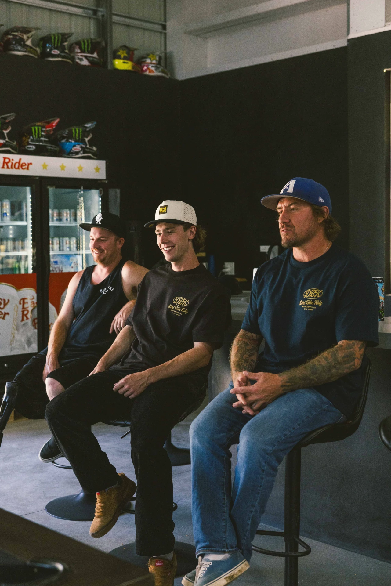 Three people seated in a shop, one wearing the DBK Gold Nugget Tee in black