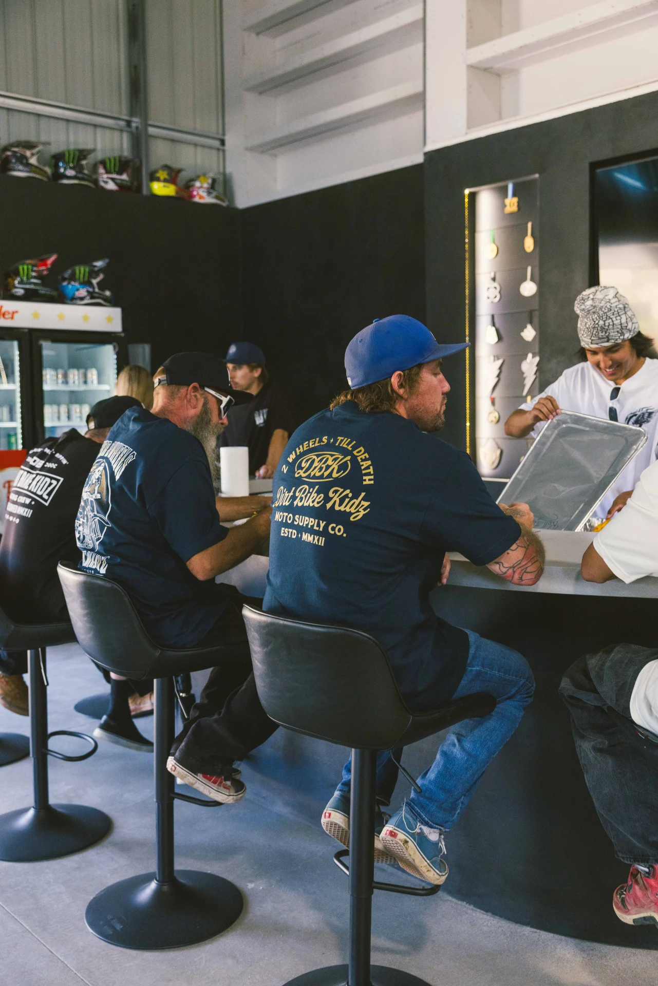 Two men seated at a shop counter, one wearing the DBK Gold Nugget Tee in navy