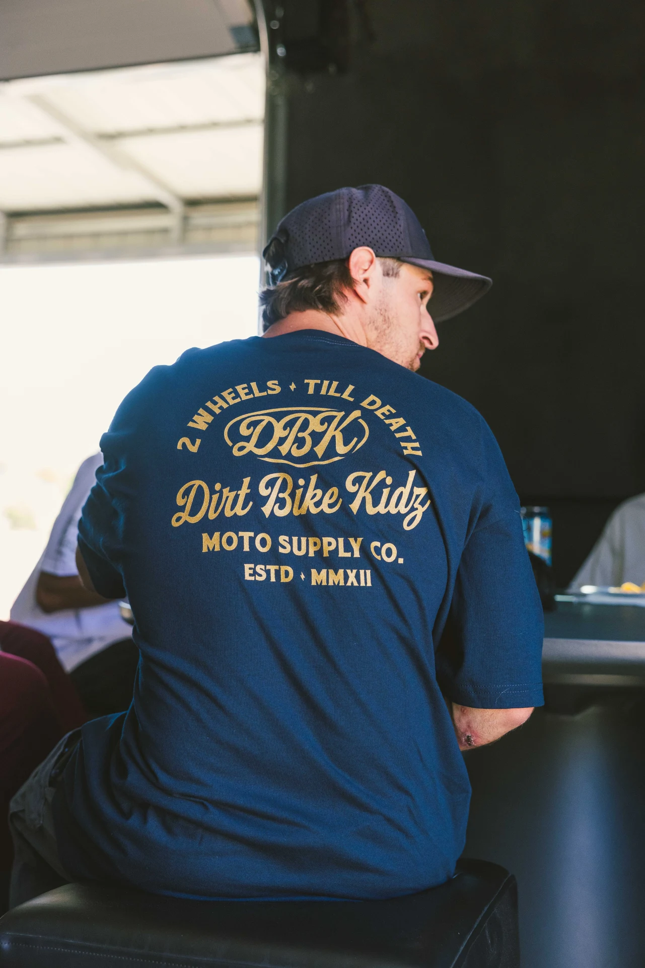 Man seated in the DBK Gold Nugget Tee in navy, back view