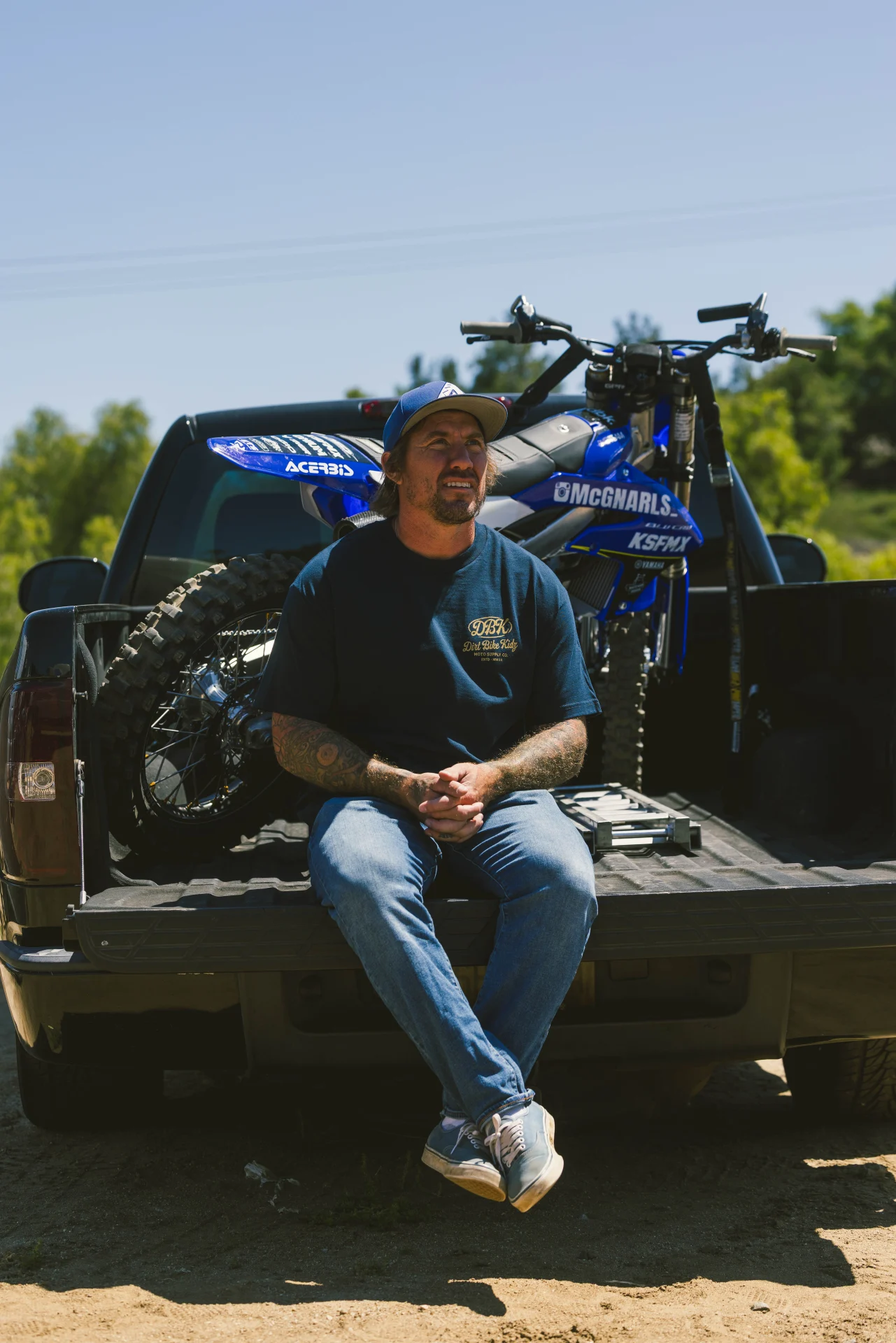 Man in the DBK Gold Nugget Tee in navy, sitting on a truck tailgate with dirt bikes behind him