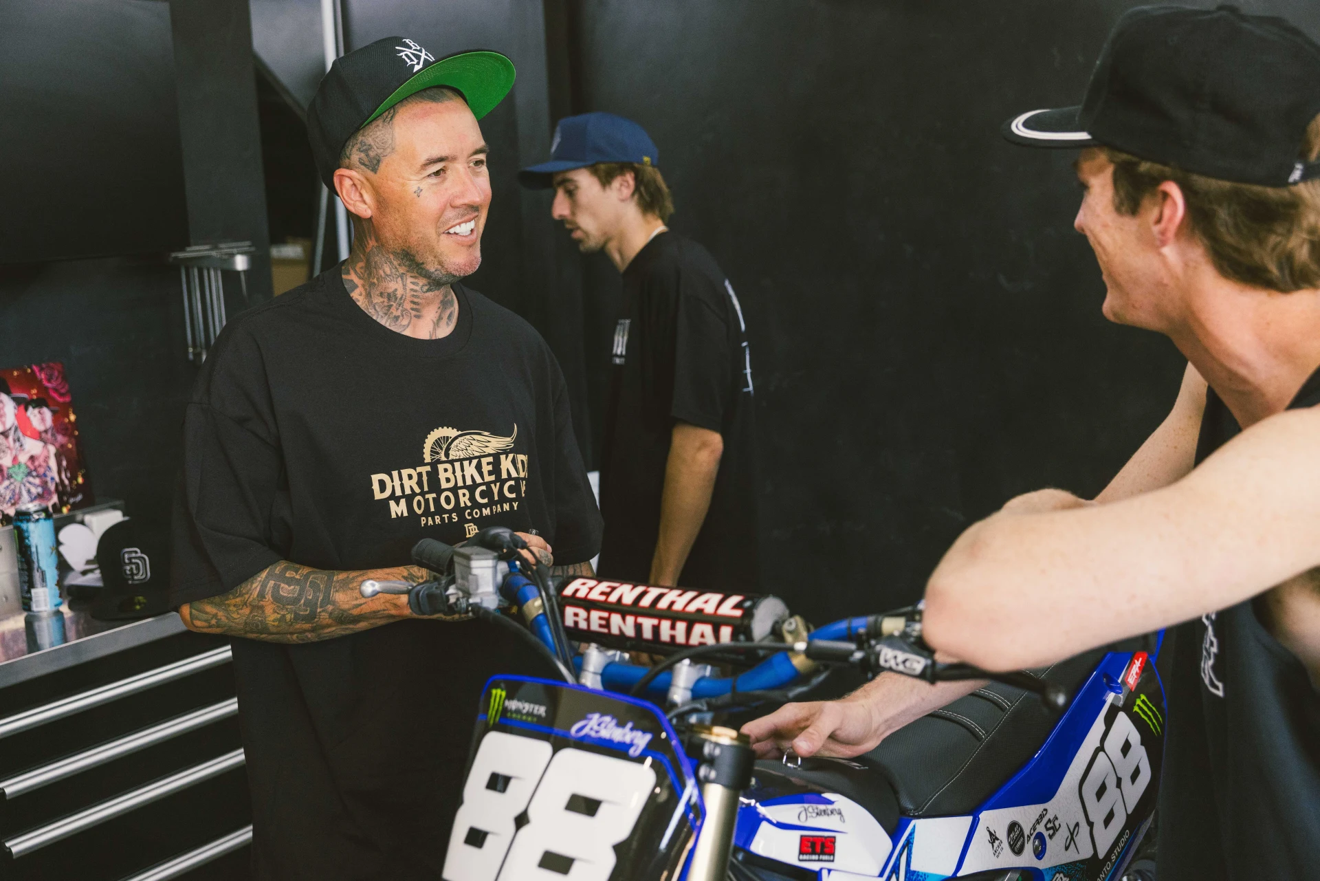 Two men talking beside a dirt bike in a shop, one wearing the DBK Gold Standard Tee in black
