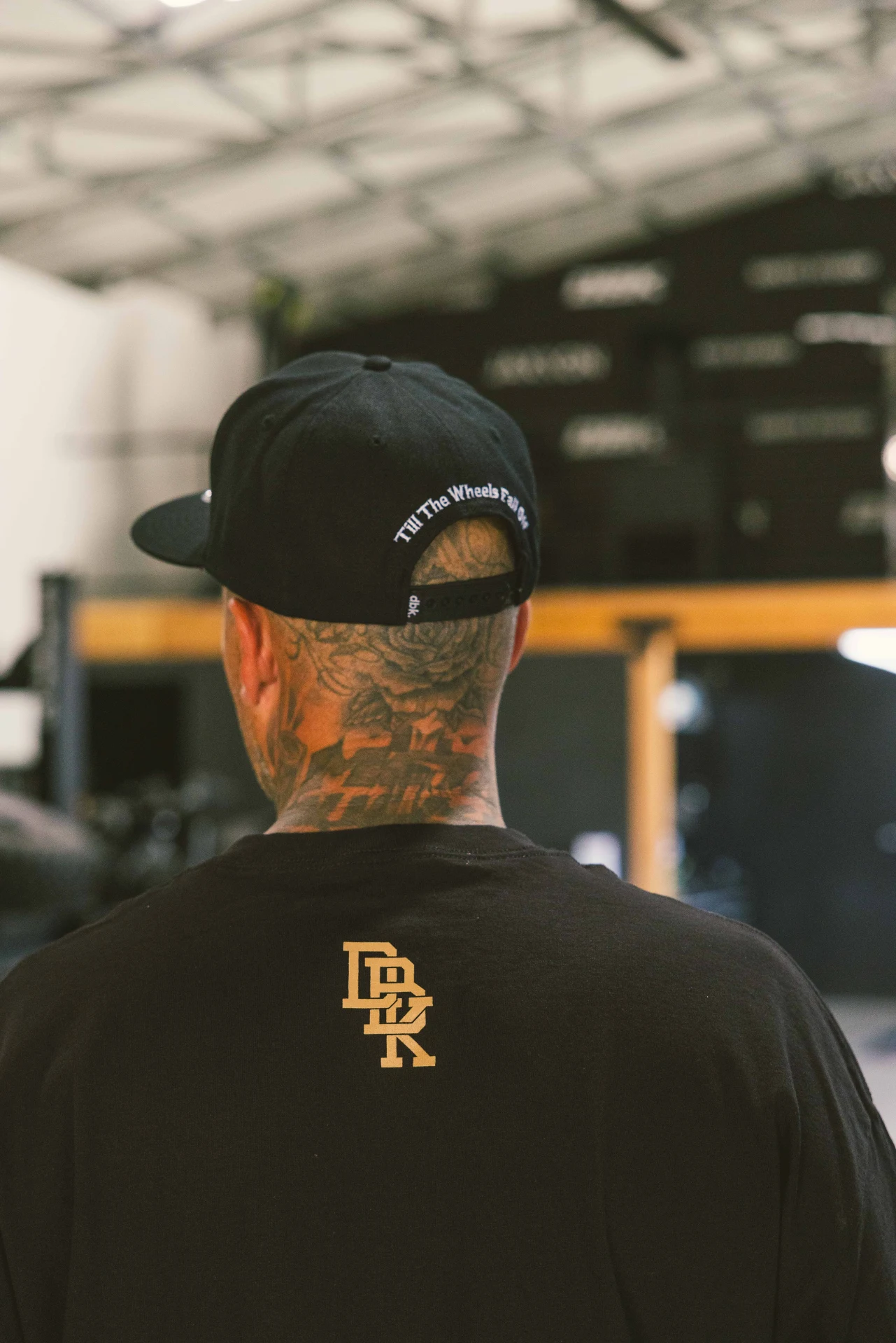Man in a cap wearing the DBK Gold Standard Tee in black, back view in a shop