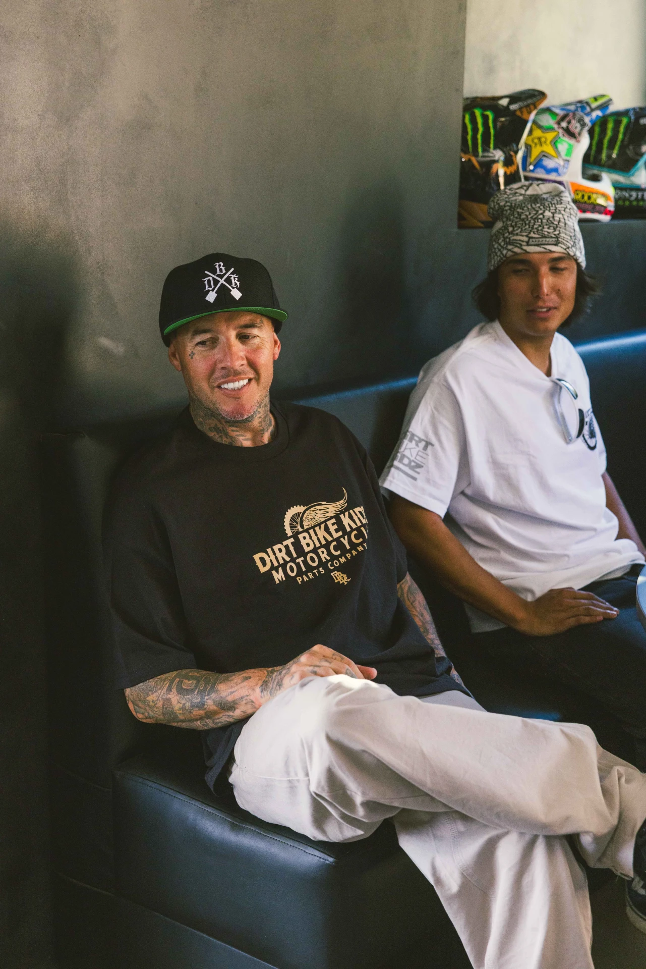 Two people seated on a couch, one wearing the DBK Gold Standard Tee in black