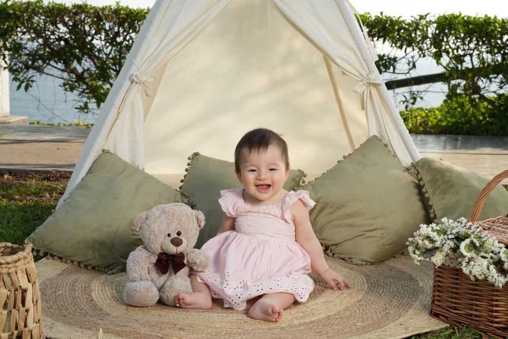 Sesión de fotos familiar en Lima: bebé con vestido rosado ríe junto a un oso de peluche frente a una carpa blanca