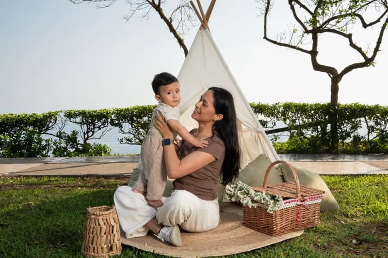 Sesión de fotos familiar en exteriores en Lima: una mamá levanta a su bebé frente a un tipi con vista al mar