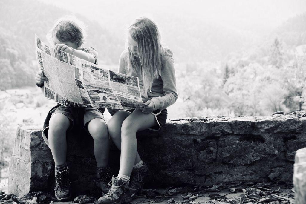 Photo of two children reading a newspaper while sitting on a stone wall