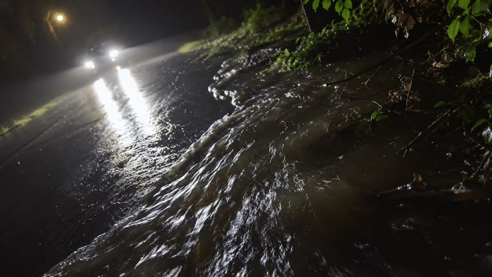 A low-angle, nighttime shot showing a road covered in flowing floodwater. The bright headlights of a car illuminate the wet, dark asphalt and the churning water moving across the surface. Lush green vegetation and tree branches are visible on the right side of the frame.