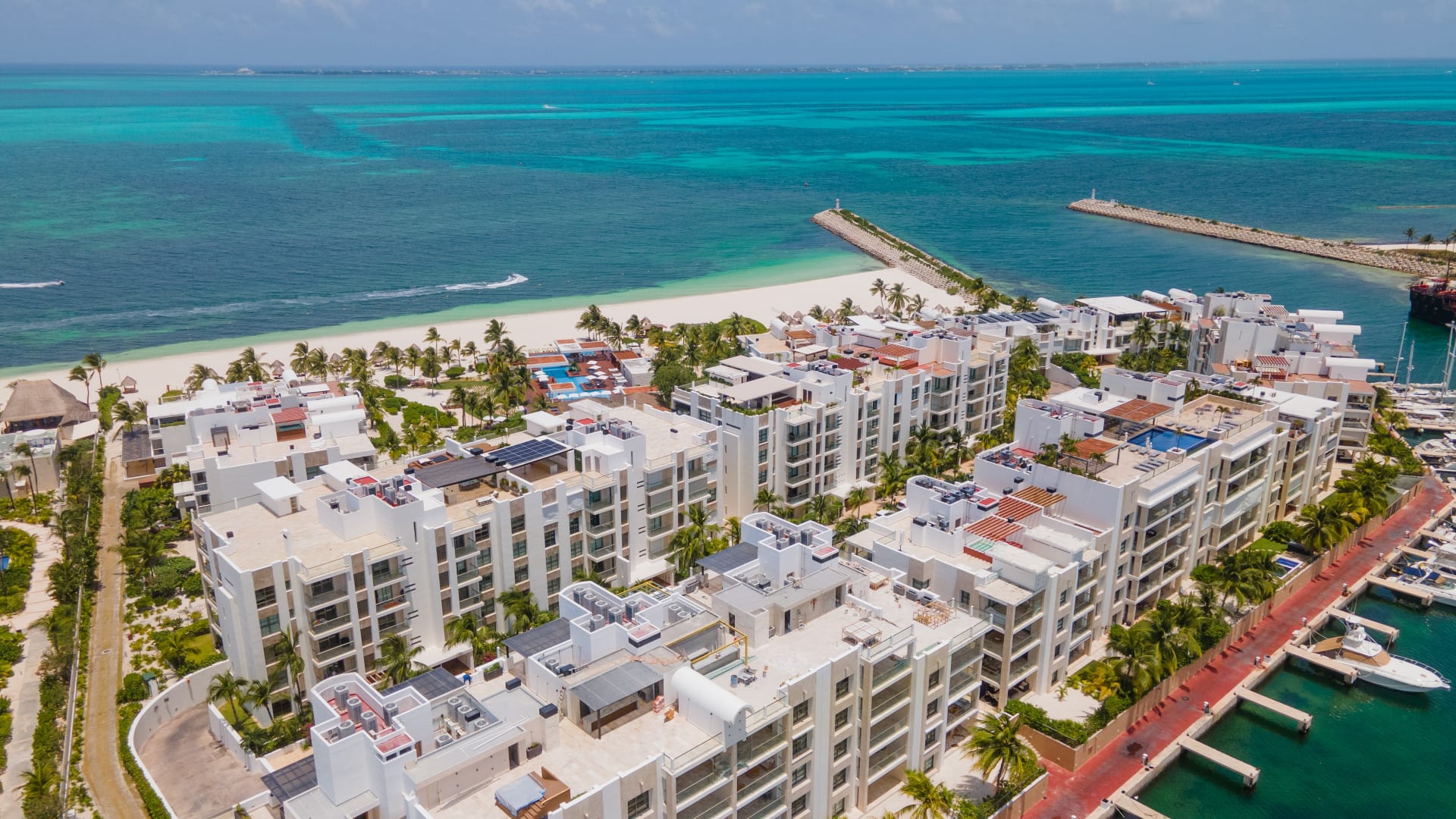 Aerial view of the La Amada residential complex in Playa Mujeres: white apartment buildings between the private beach and the marina, with the Caribbean beyond.