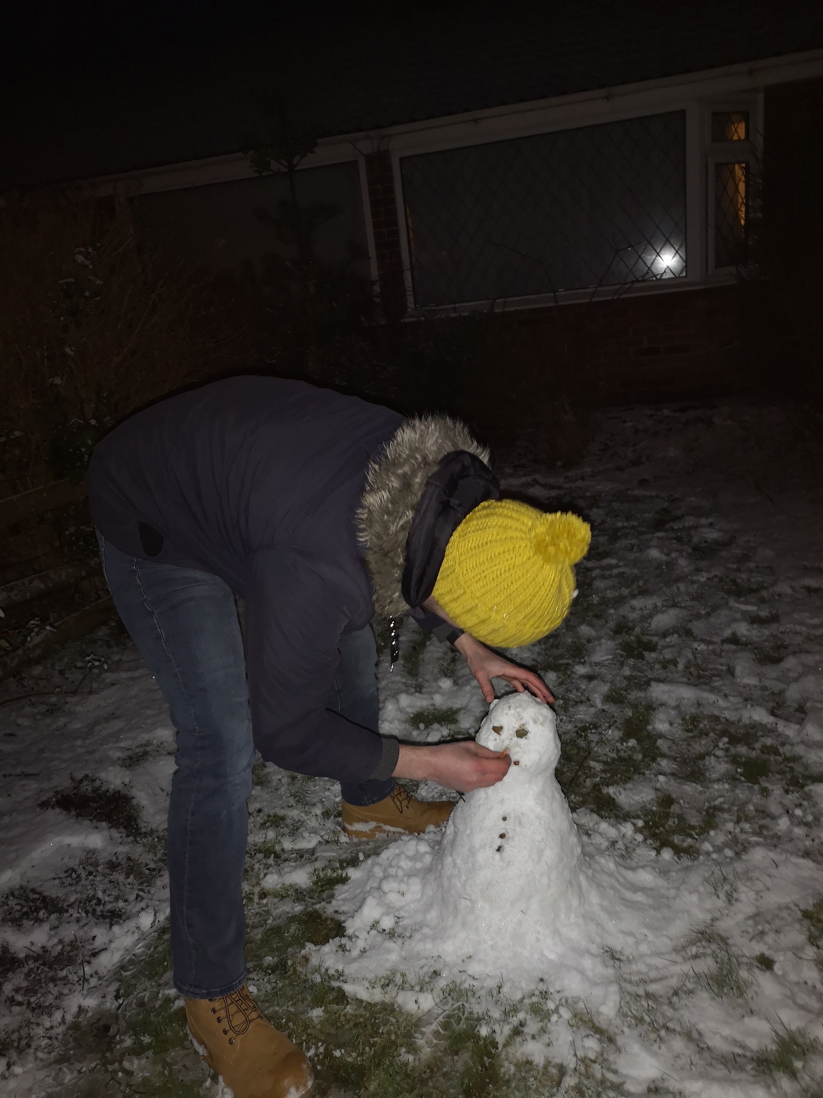 Joe in a padded navy coat with a bright yellow crocheted hat on, bending down to put a carrot nose onto a small snowperson which is as tall as Joe’s knees. There’s a dusting of snow on the ground, with the flash of the camera reflected in the glass of the bungalow in the background. It’s very dark, with the bright white snow of the snowperson providing a stark contrast against the dark background.