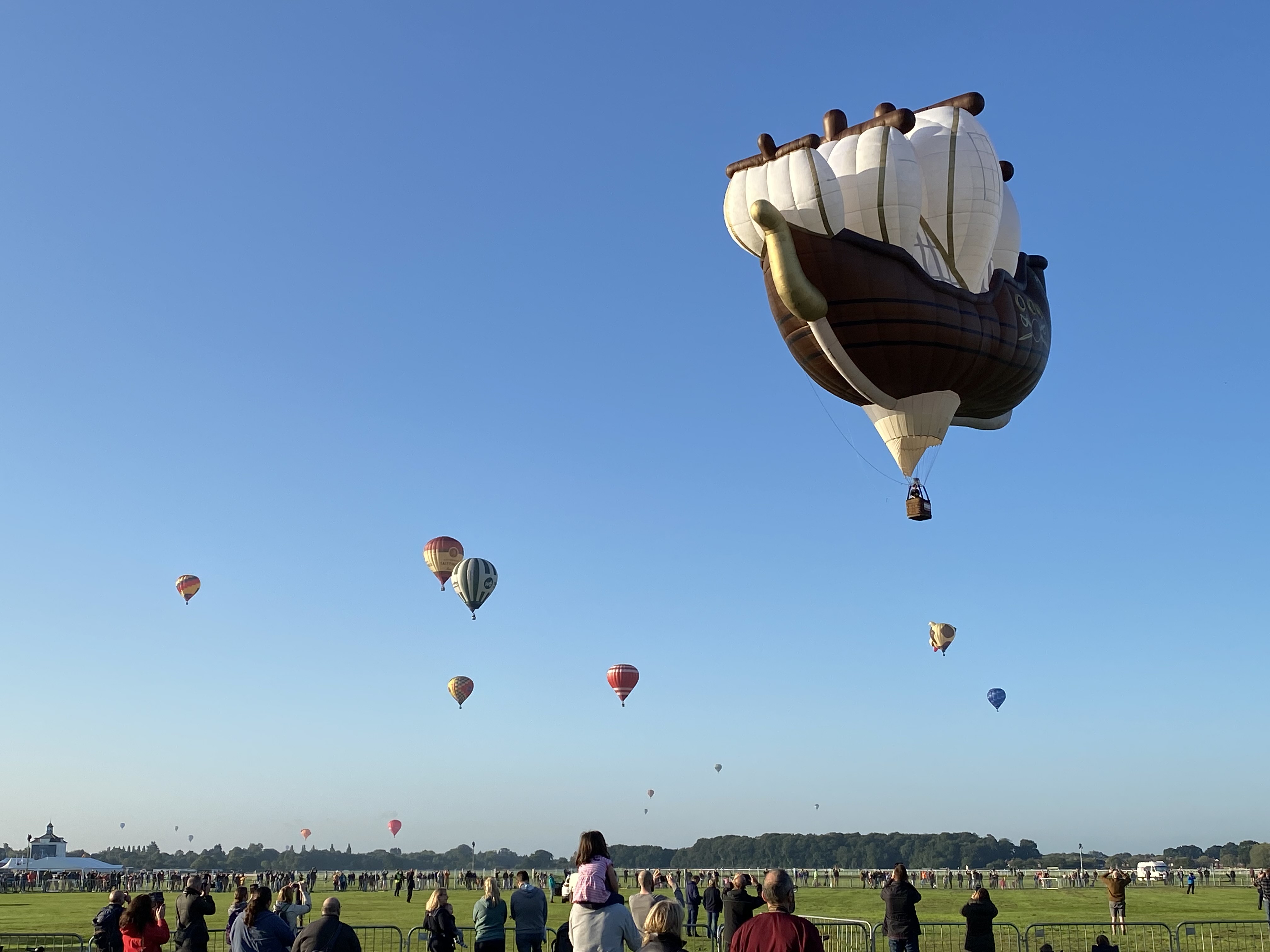 A pirate ship hot air balloon is present in the foreground of the image with a series of other different coloured hot air balloons visible across the clear blue sky in the background. At the bottom of the image, crowds of people are stood behind barriers watching the pirate ship balloon taking off into the sky.