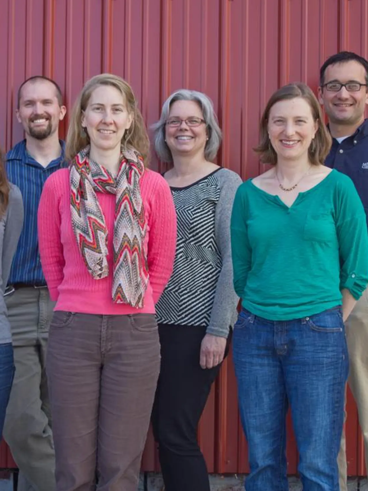 Seven people stand smiling in front of a red wall.
