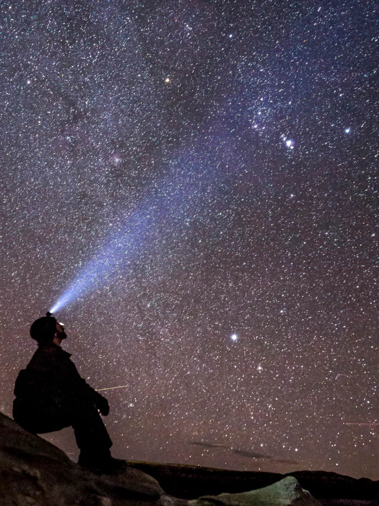 Someone sits on a rock wearing a helmet with a light shining up at a dark sky full of stars.