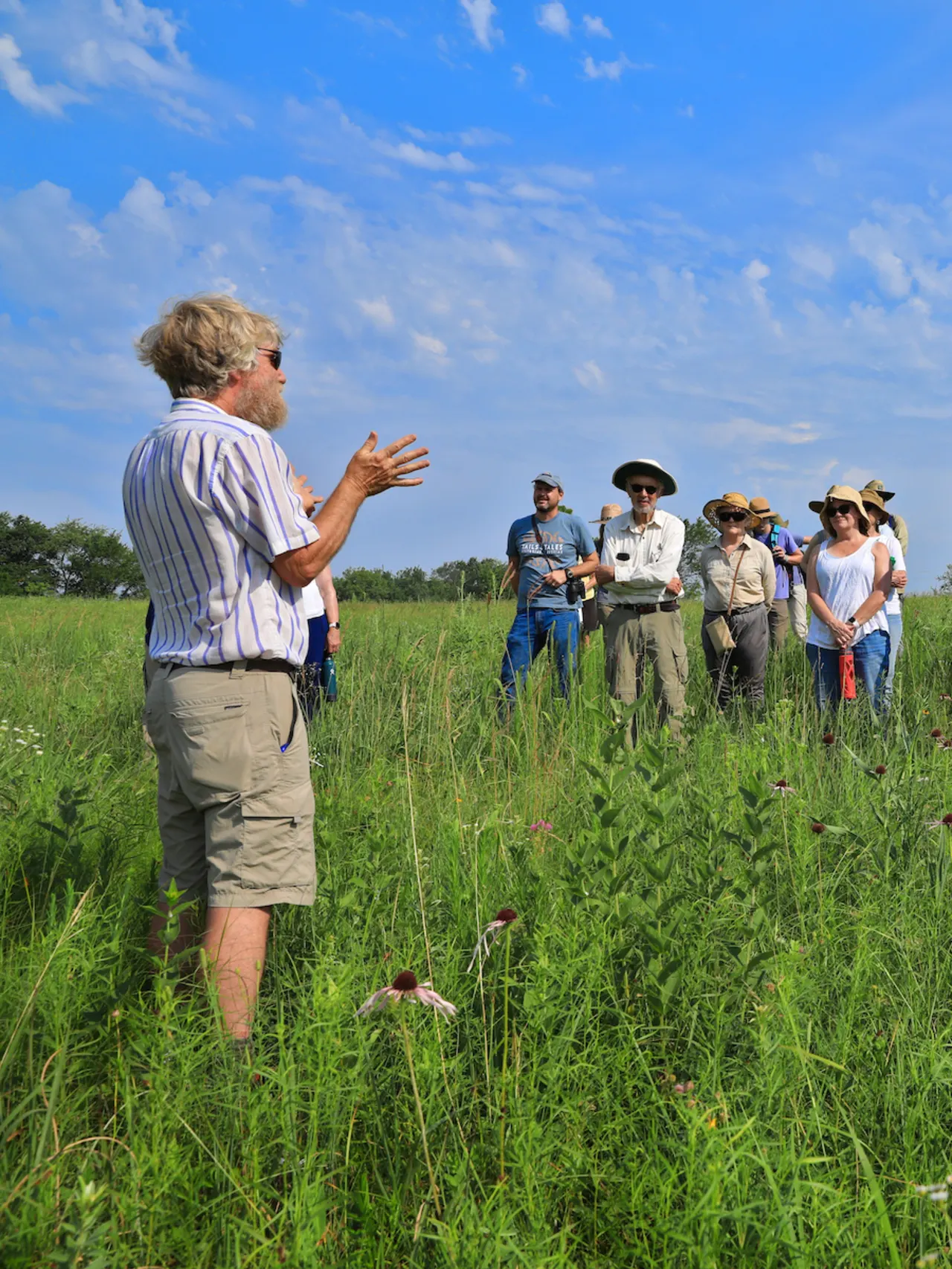 A group of people standing in a field with flowers looking at one person who is talking.