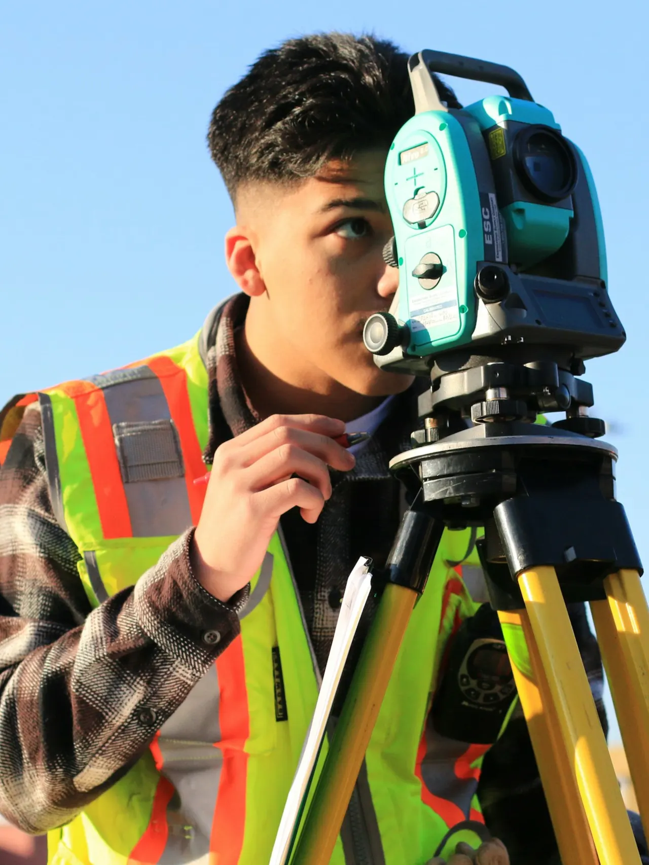 Someone wearing a reflective vest standing behind a camera and looking into its lens with one eye