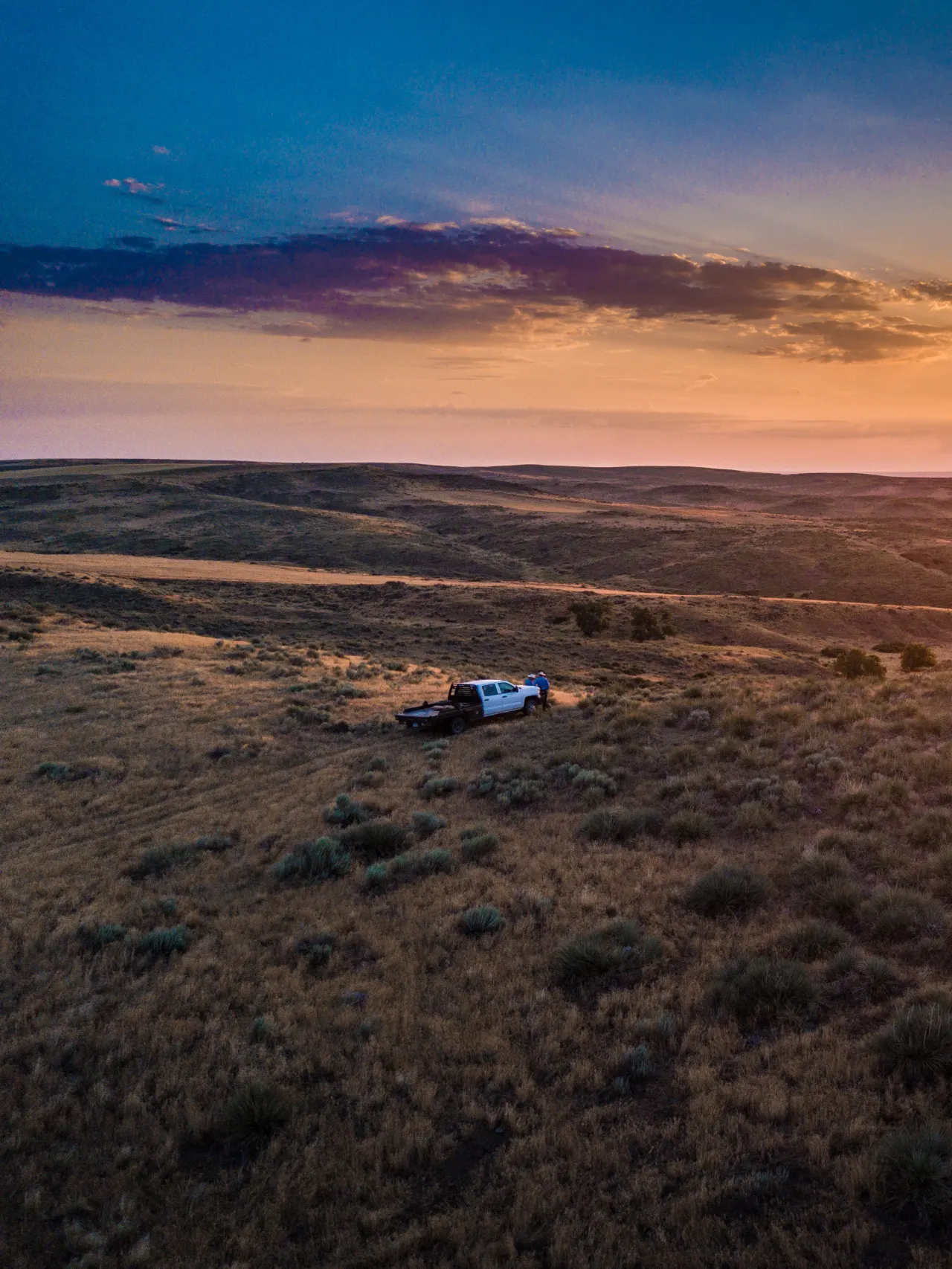 Photo of a sun setting over rolling hills, with a white truck with people leaning on it looking at the sunset.