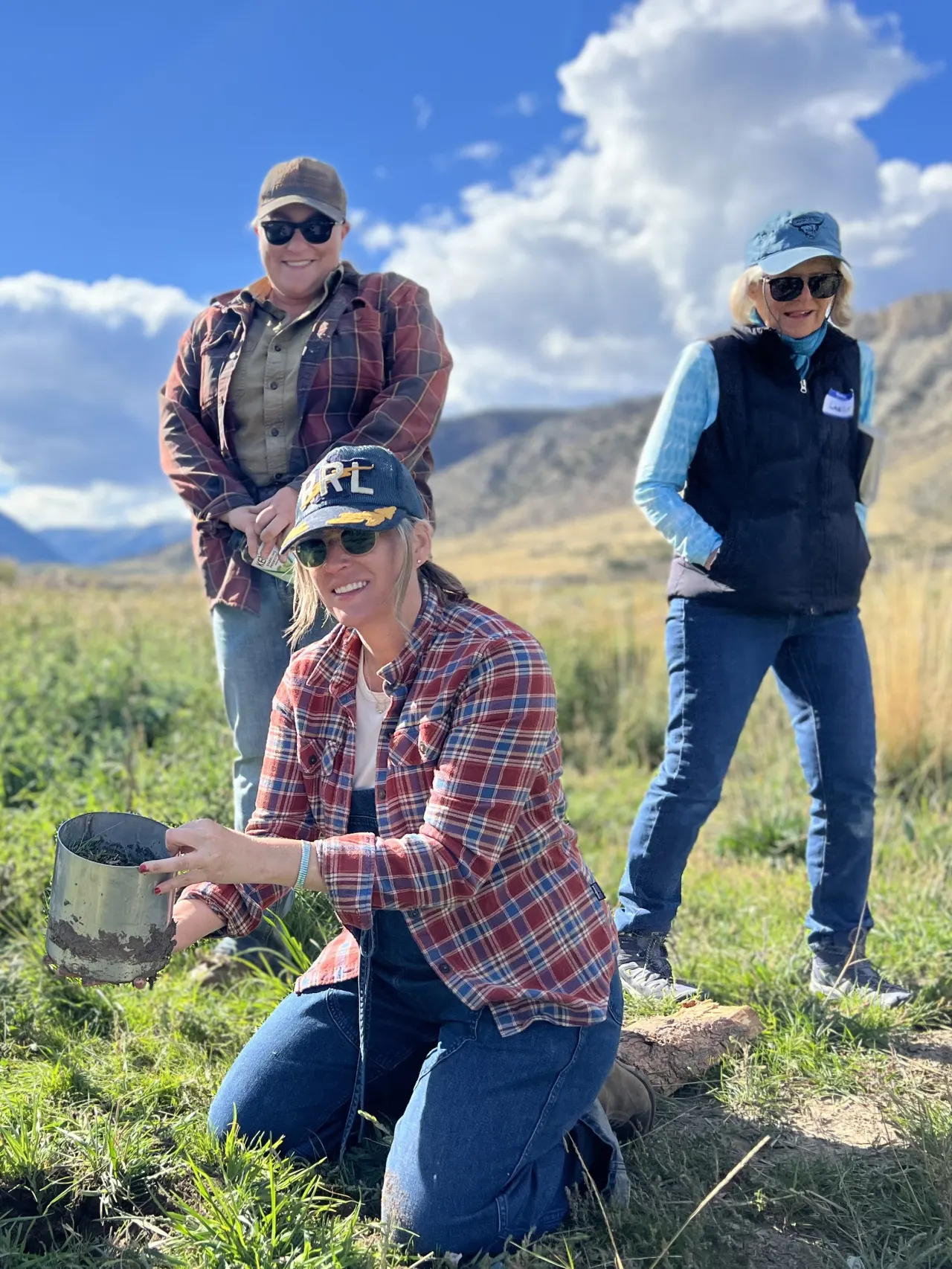 Four women in a field wearing hats and working gear, one holding a bucket and one with a shovel.