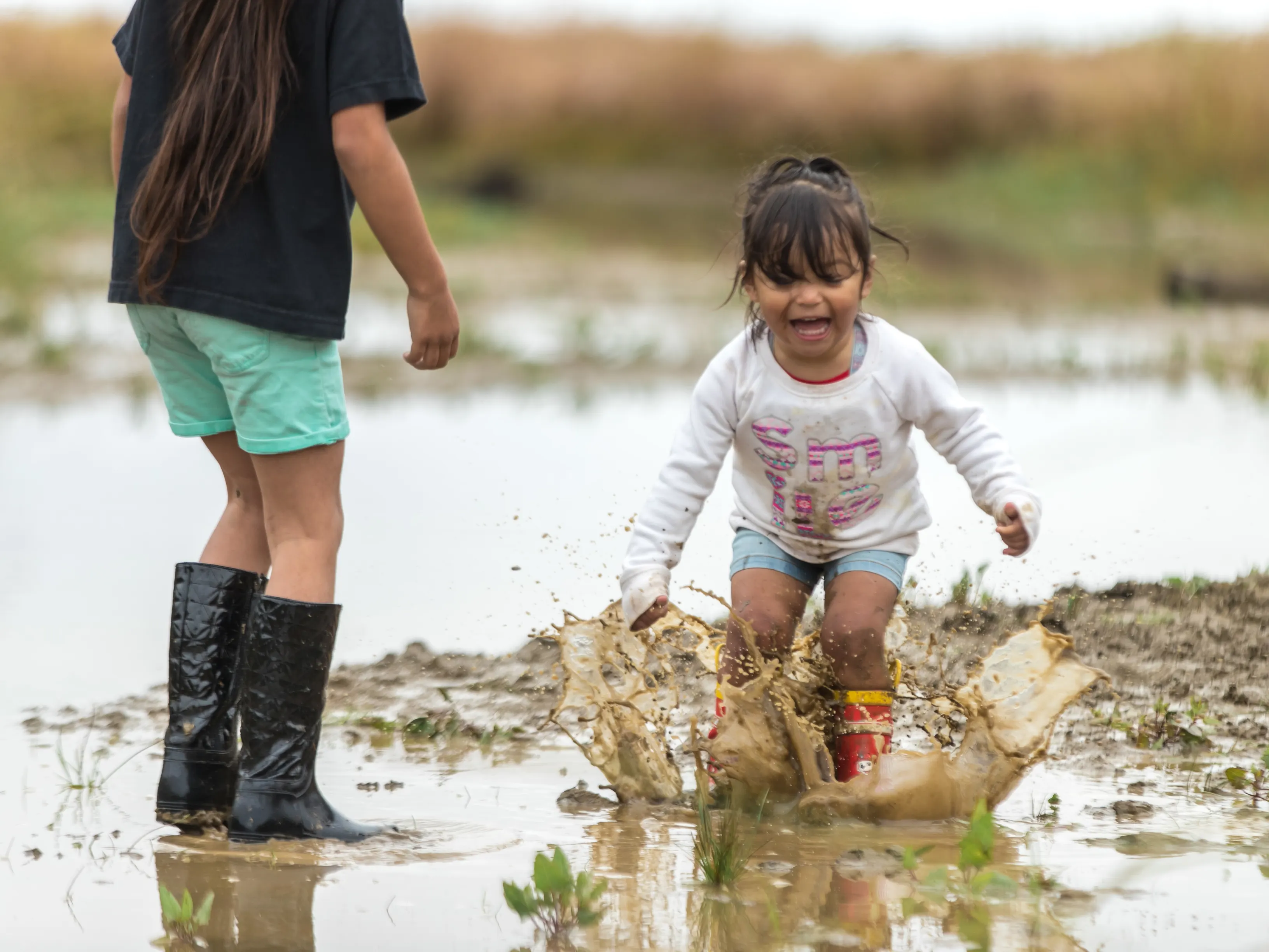 Little girl wearing rainboots jumping in a puddle of mud laughing