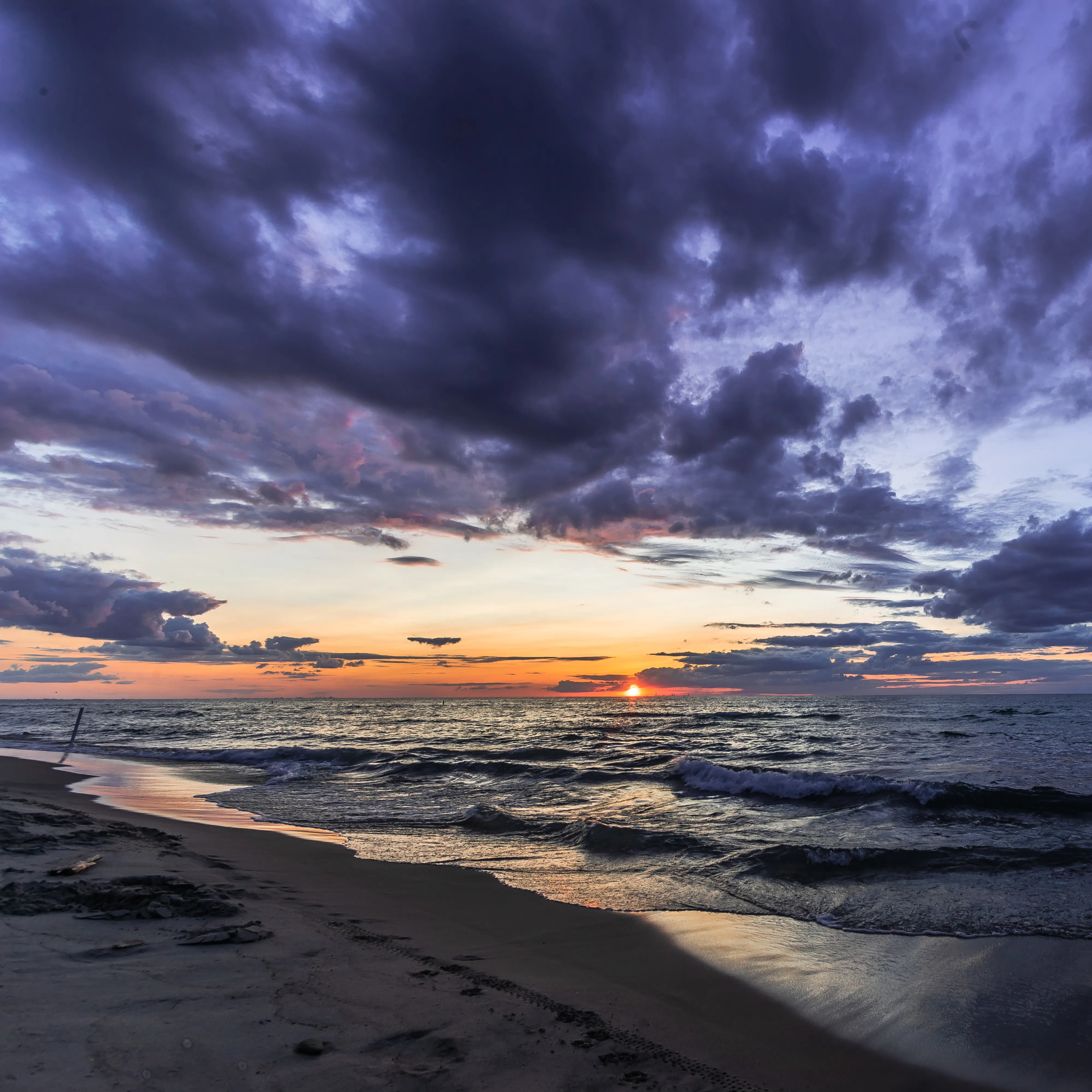 Photo taken on a beach overlooking the waves, with the sun setting across the ocean. Dark clouds are overhead.