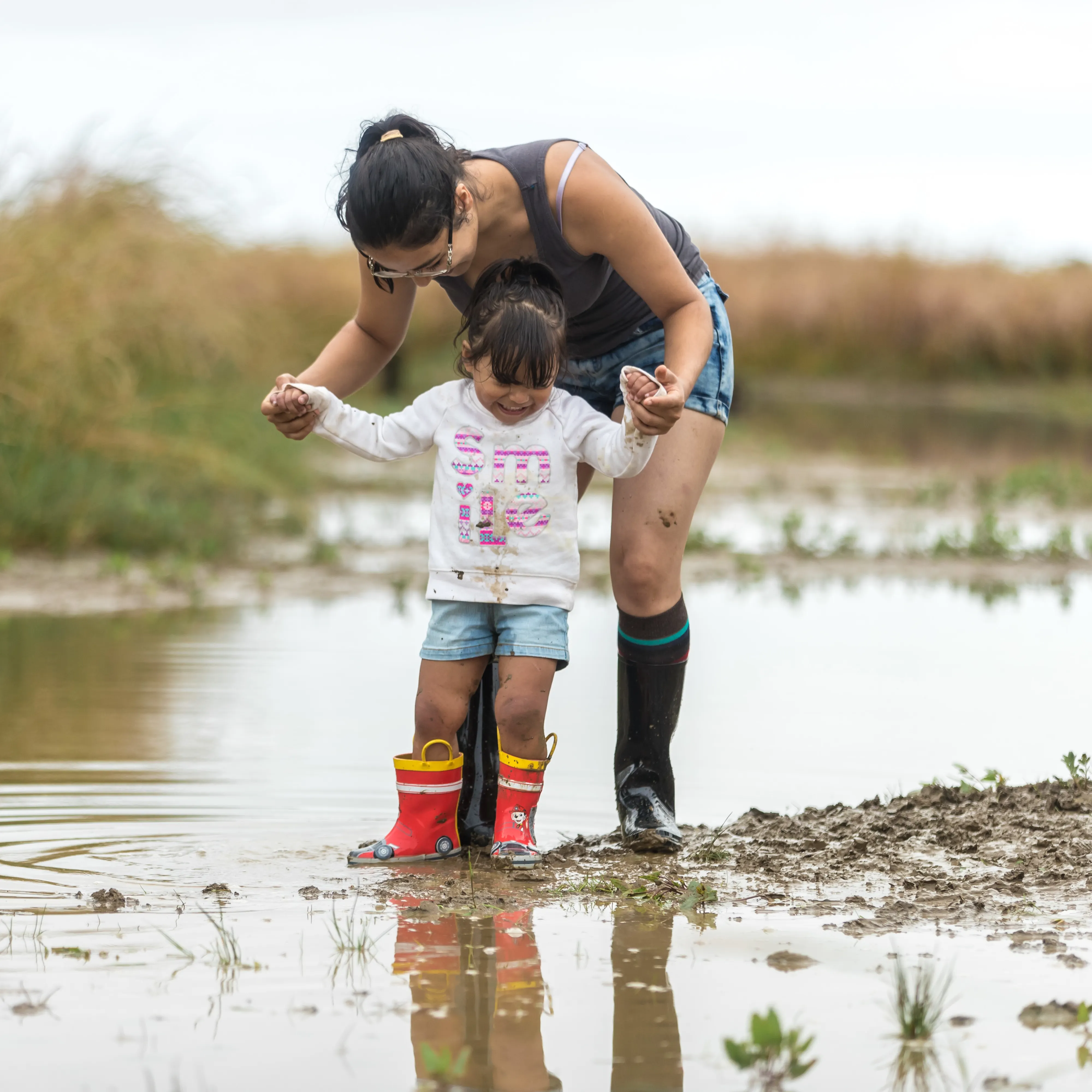 A child standing a puddle of muddy water wearing rain boots and smiling widely with a woman also in boots standing above her holding her hands.