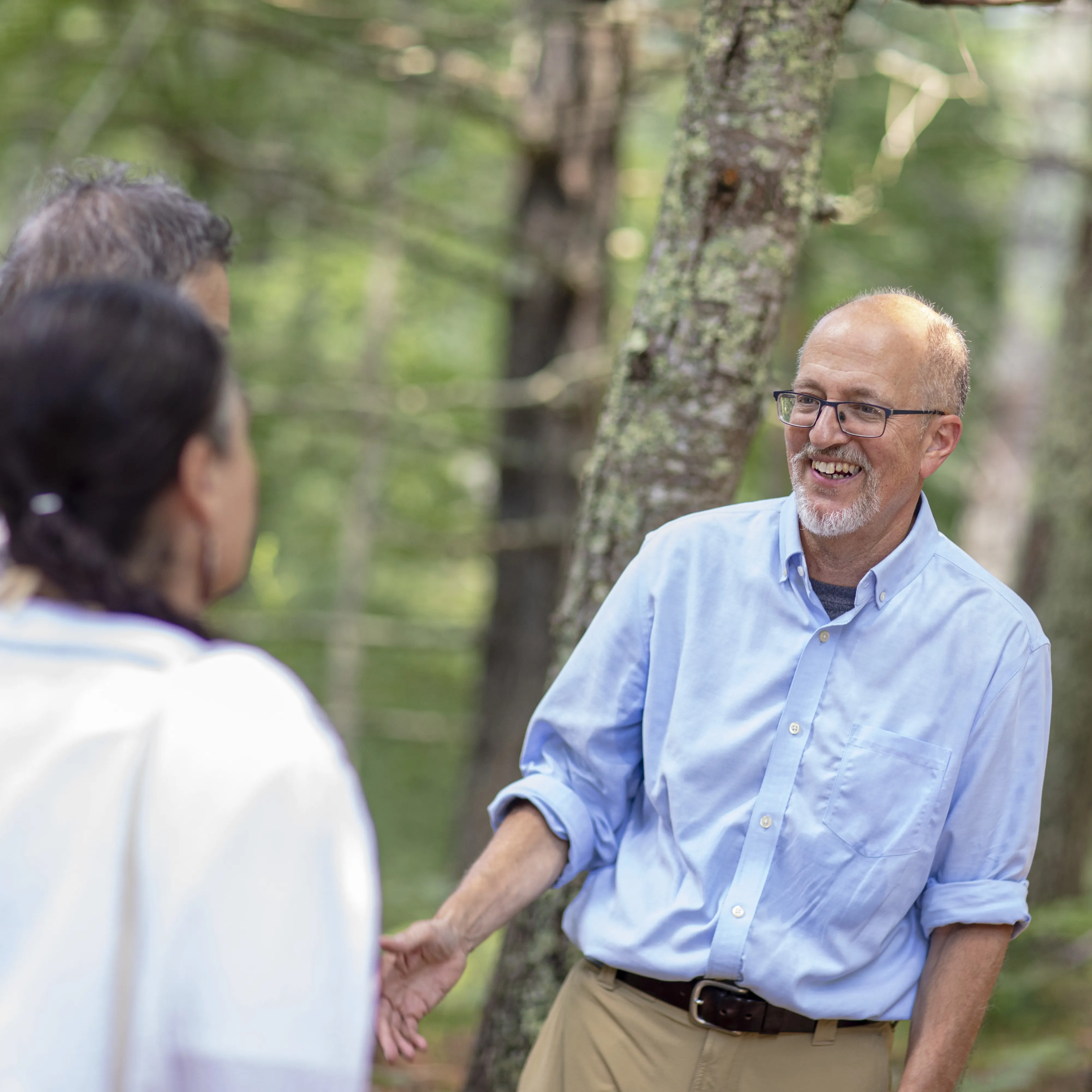 A man wearing glasses and a blue shirt is laughing while talking to two people who have their backs turned to the camera.