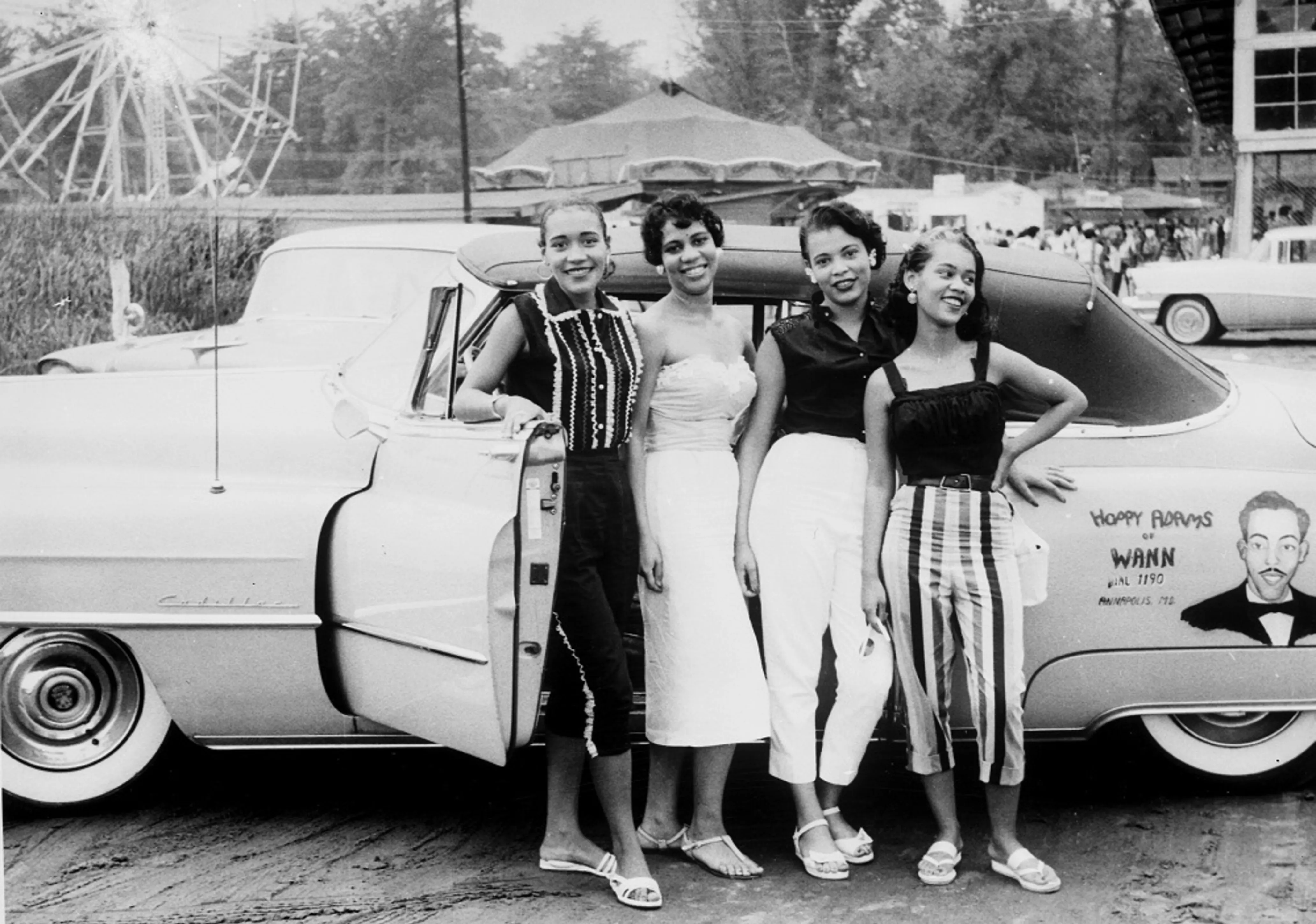 Four Black young women stand in front of a car parked at the beach