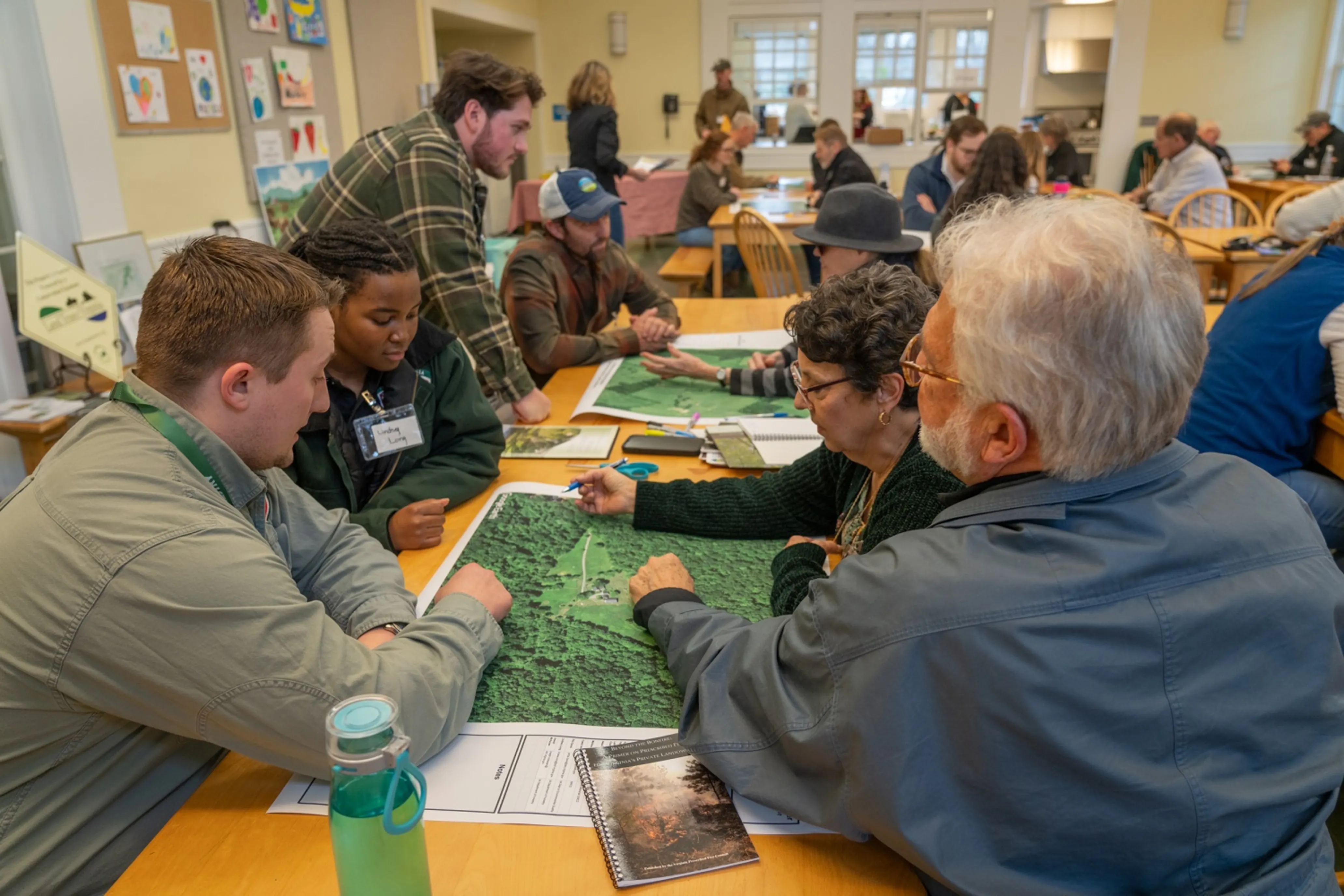 People in a room gather around tables, discussing and examining green maps, with papers and notebooks scattered around.