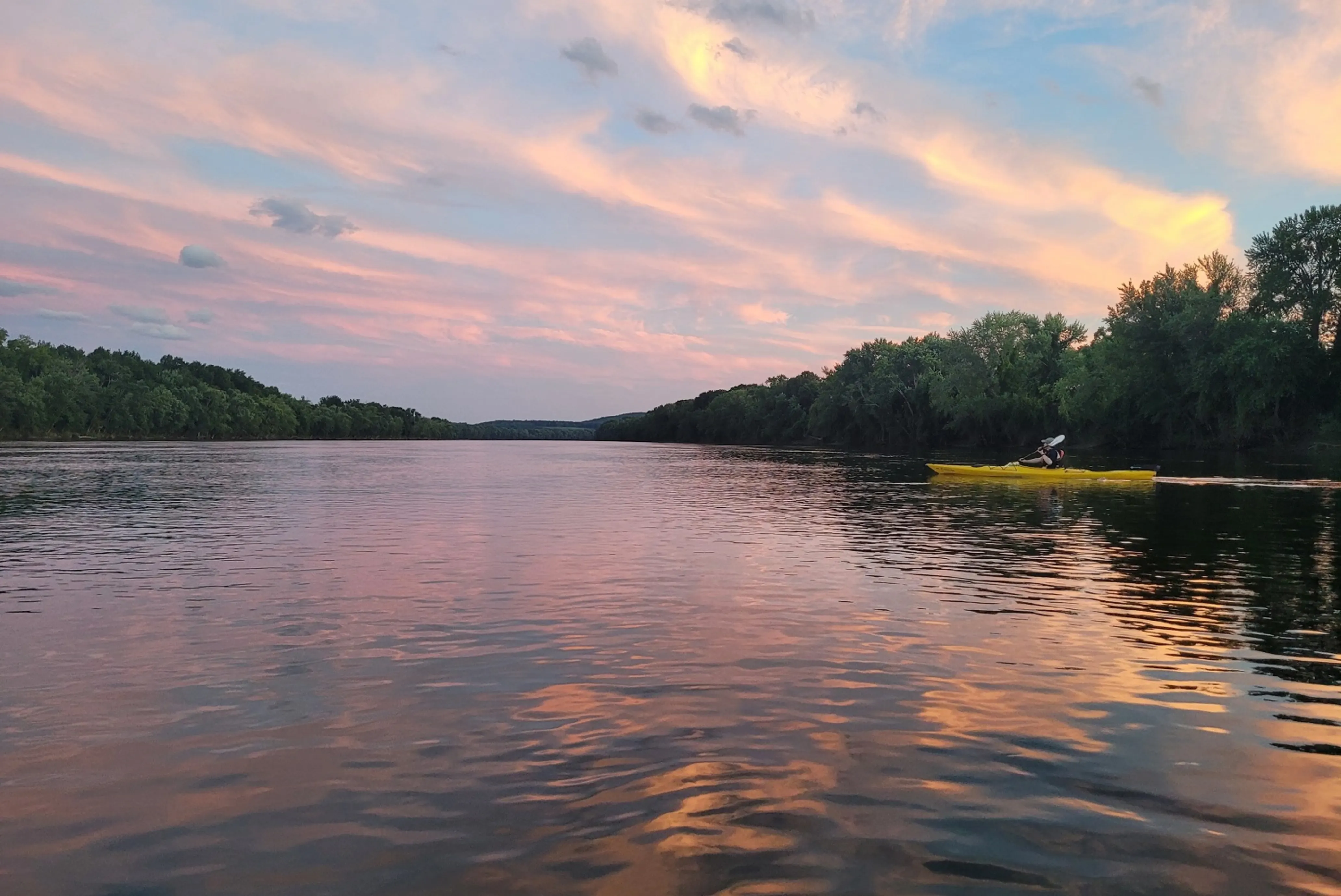 A person kayaks on a calm river at sunset, with pink and orange clouds reflecting on the water and trees lining the riverbank.