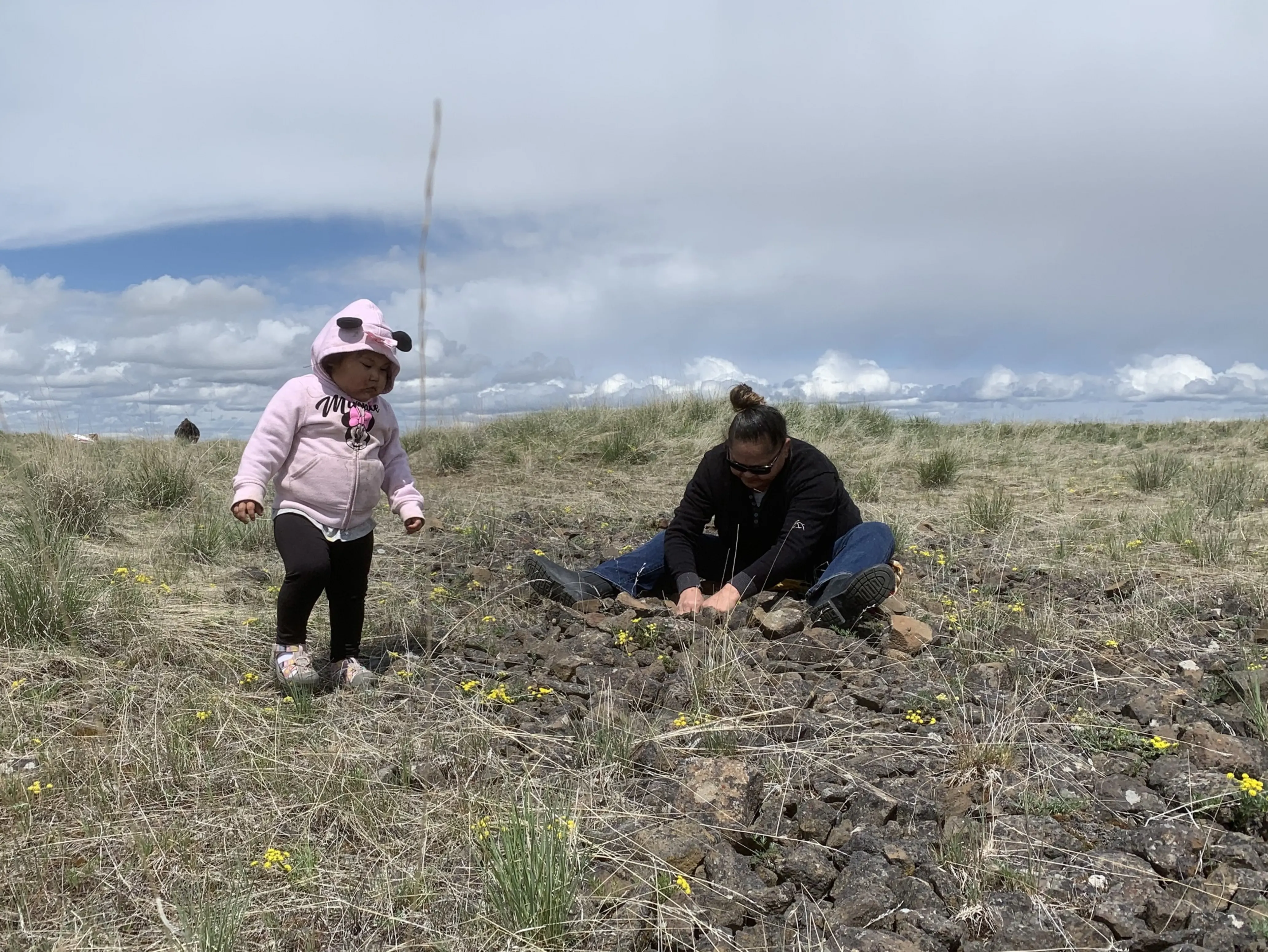 A mother sits on the ground with her young daughter standing nearyby, picking roots on a stretch of open grassland.