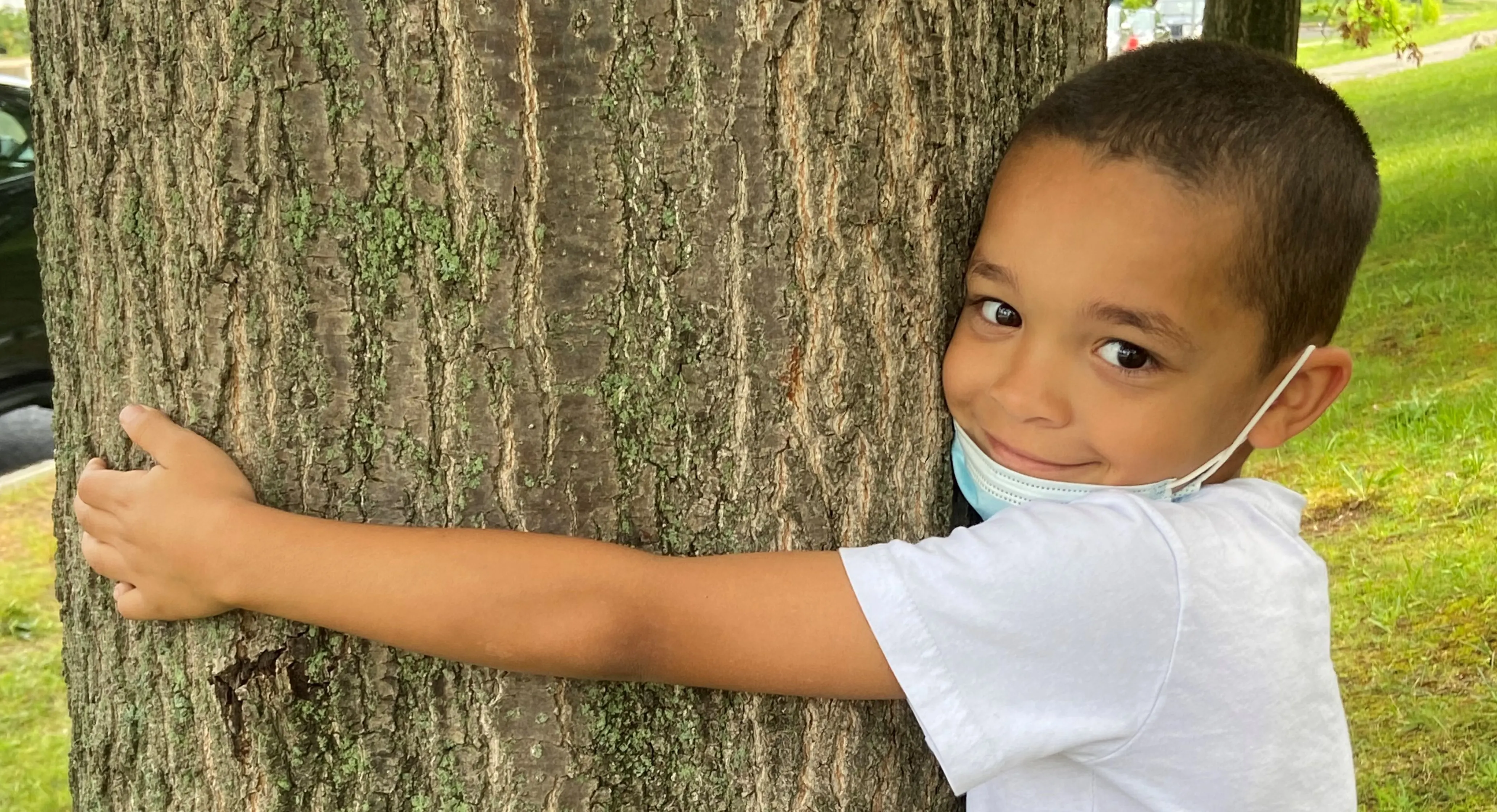 Young black child who appears to be male wearing a mask pulled down below mouth hugging a tree.