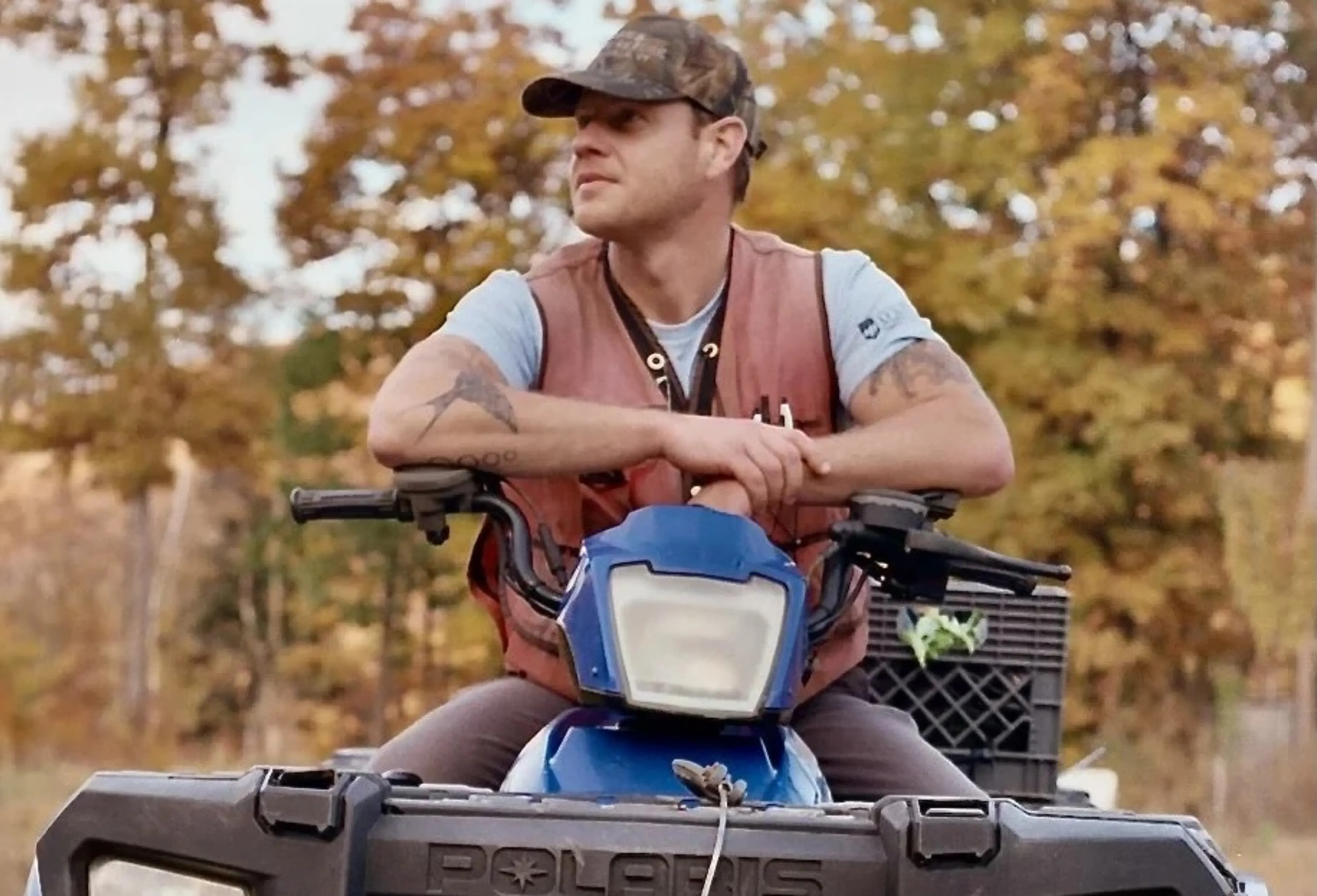 Man sitting on a blue ATV, wearing a camo cap and brown vest, in a wooded area with autumn foliage.