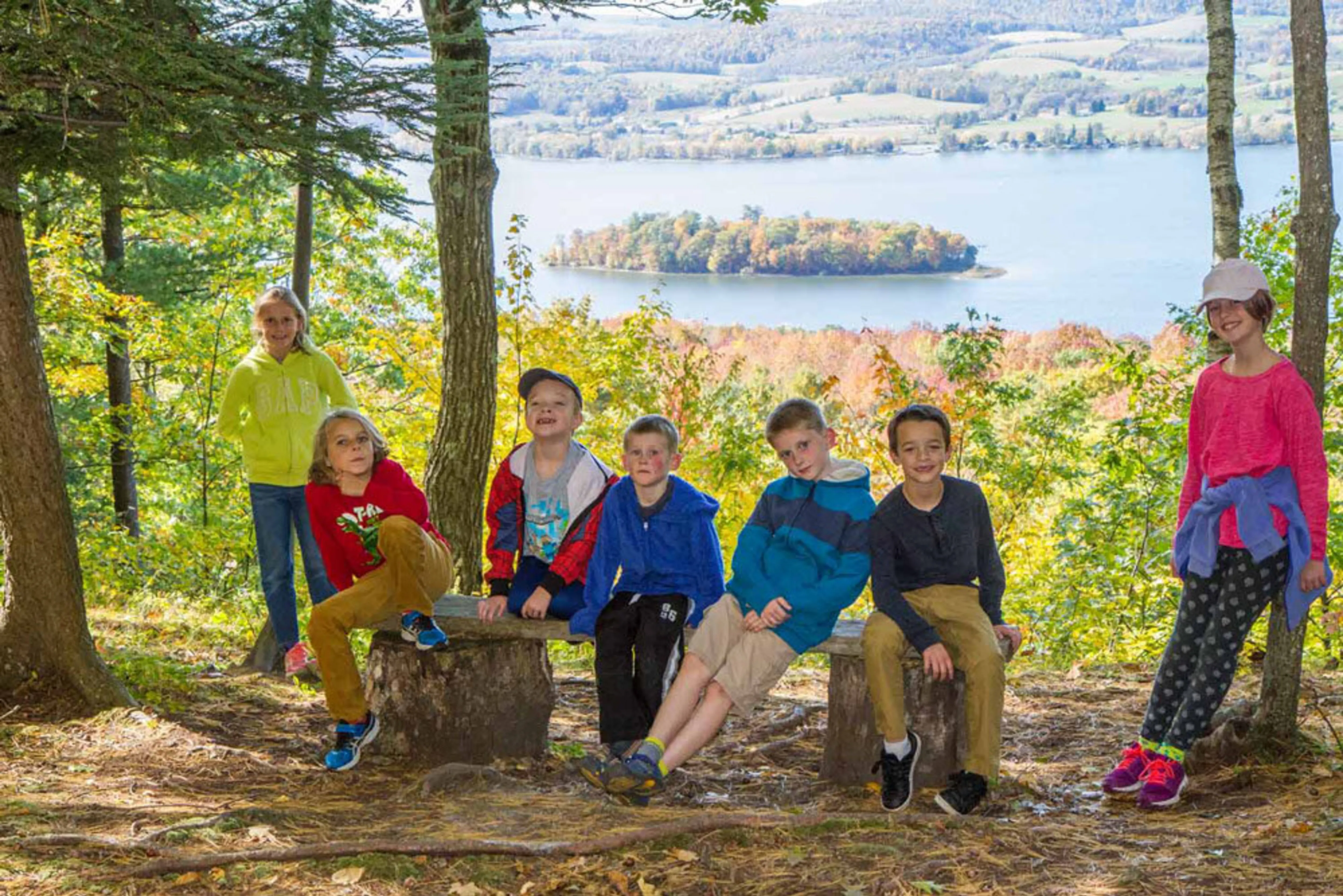 Children around 7-10 years old sitting on a bench made out of tree trunks in the woods, with a river behind them and an island filled with trees in the river.