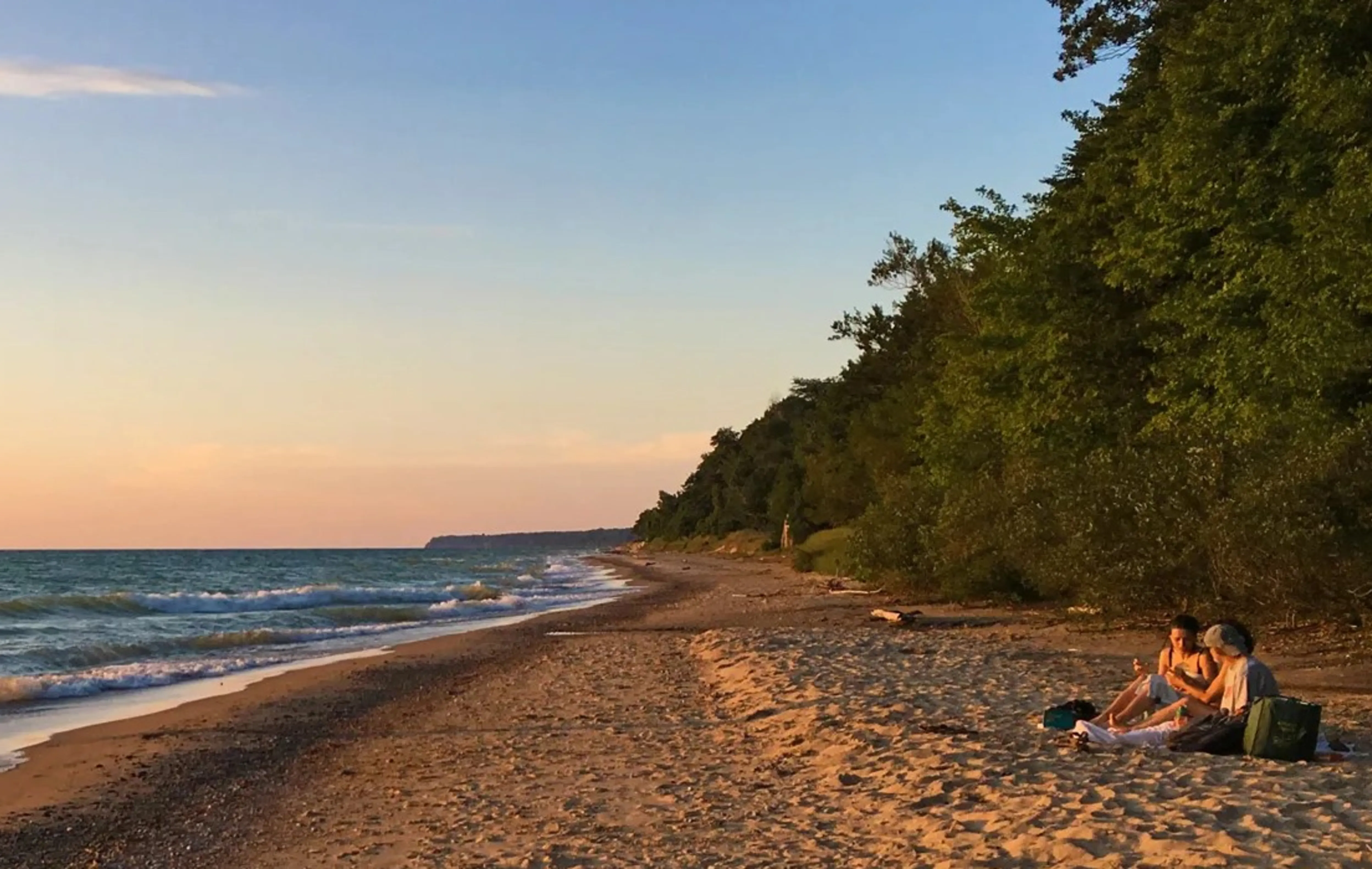 Two people sitting on a sandy beach eating a picnic with water on one side and trees on the other