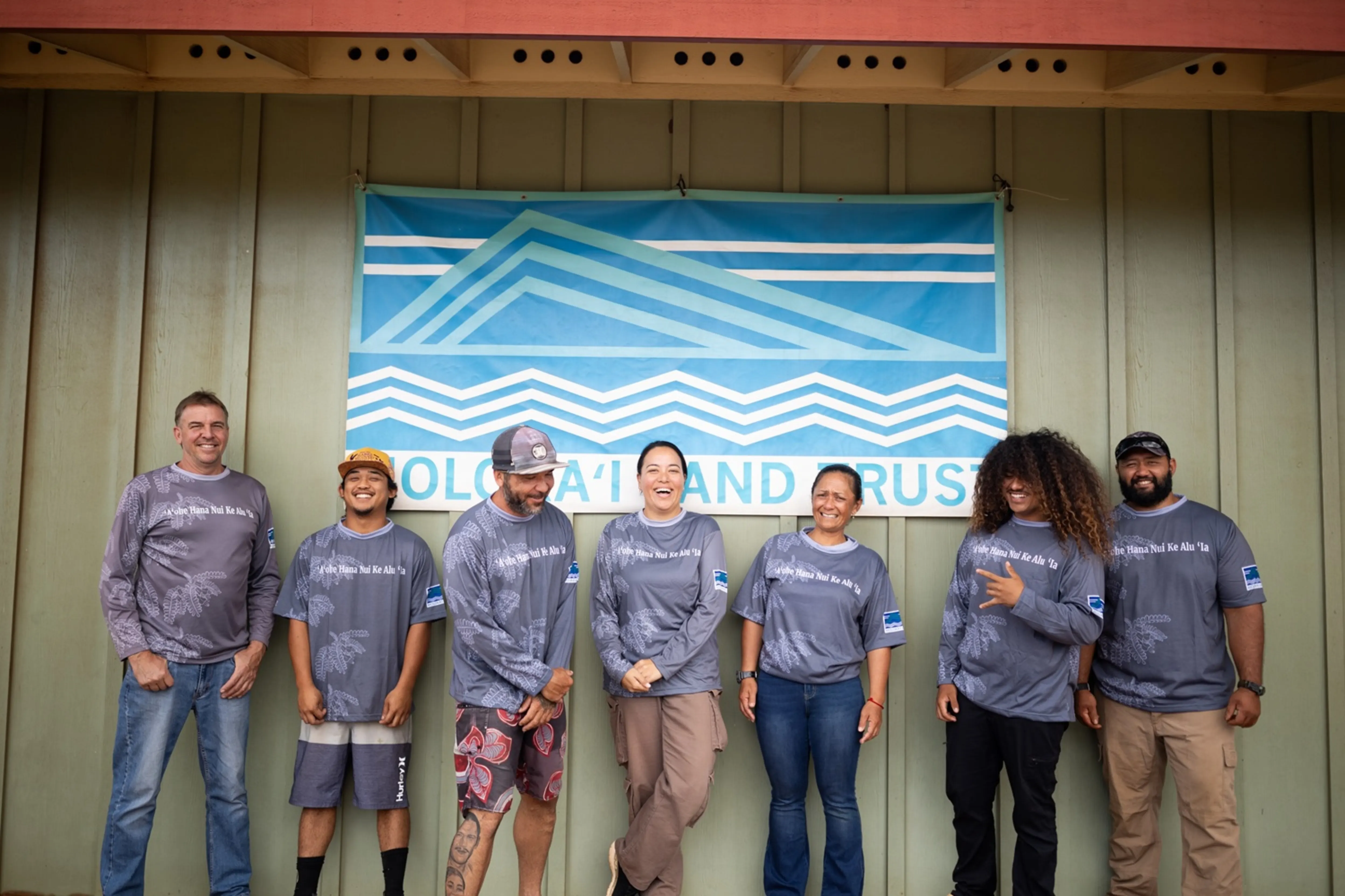 Seven people in matching gray shirts stand smiling in front of a "Molokaʻi Land Trust" banner on a wood-paneled building.