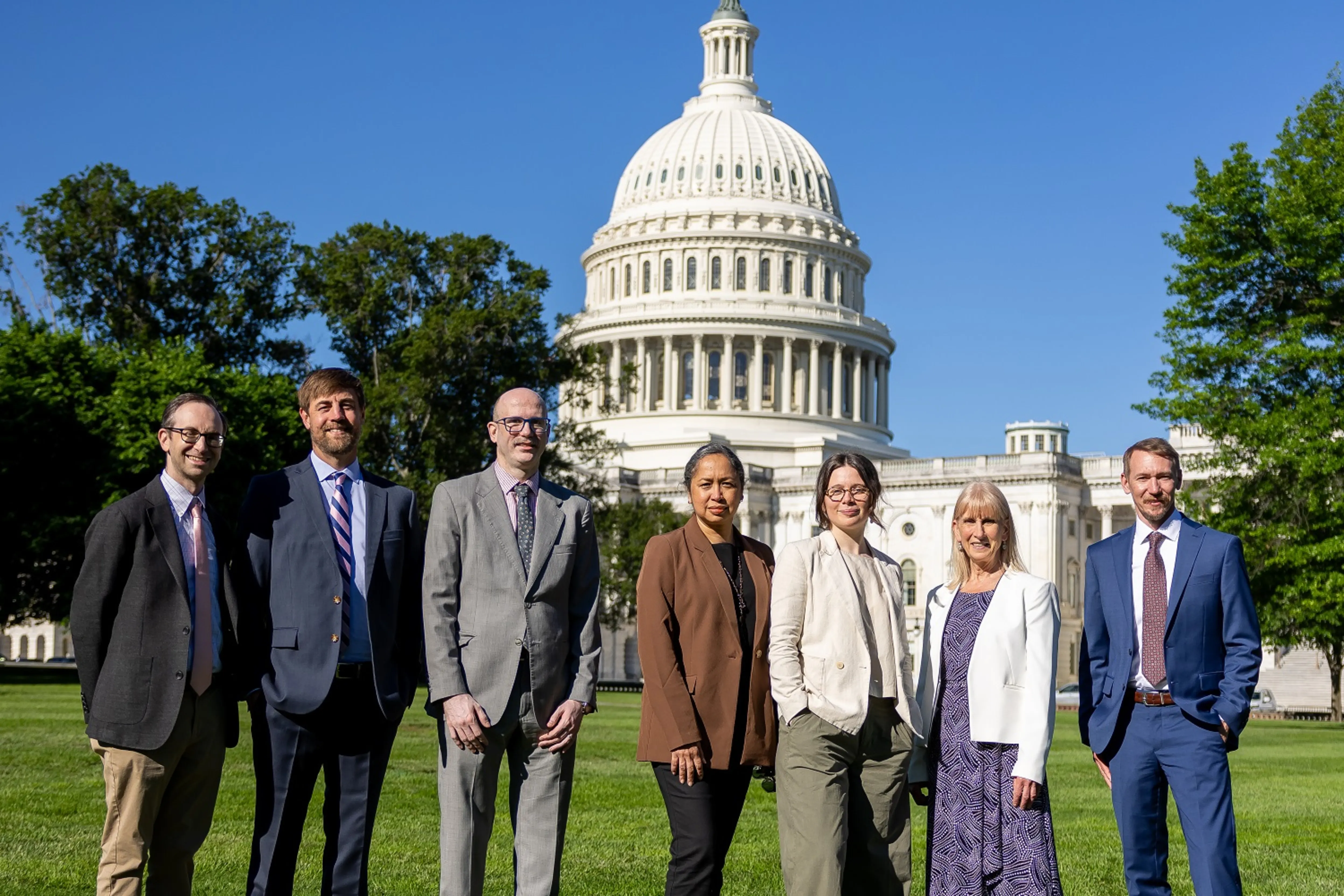 Seven professionally dressed people posing on the lawn with the U.S. Capitol dome in the background.